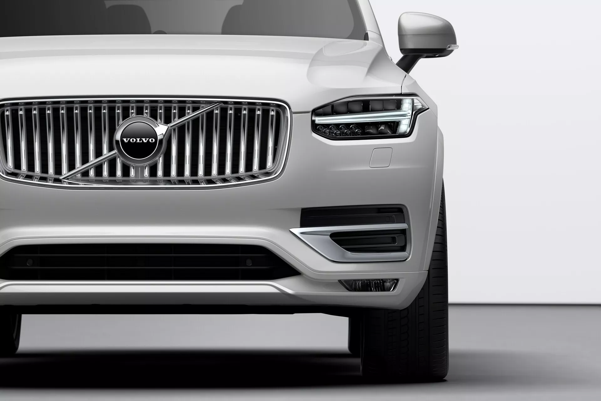 Imagen en primer plano de la parte delantera de un Volvo XC90 plateado de 2015 (2da generación) tomada desde un ángulo bajo. Destacan la rejilla cromada prominente con el emblemático logo de Volvo, los elegantes faros con luces diurnas LED en forma de T, conocidas como 'Martillo de Thor', y los sensores de aparcamiento bien integrados en el parachoques delantero. La parte inferior del parachoques incluye tomas de aire y alojamientos para los faros antiniebla bordeados por un adorno plateado. Los espejos exteriores visibles sugieren características tecnológicas avanzadas, como indicadores de dirección o sistemas de advertencia de punto ciego.