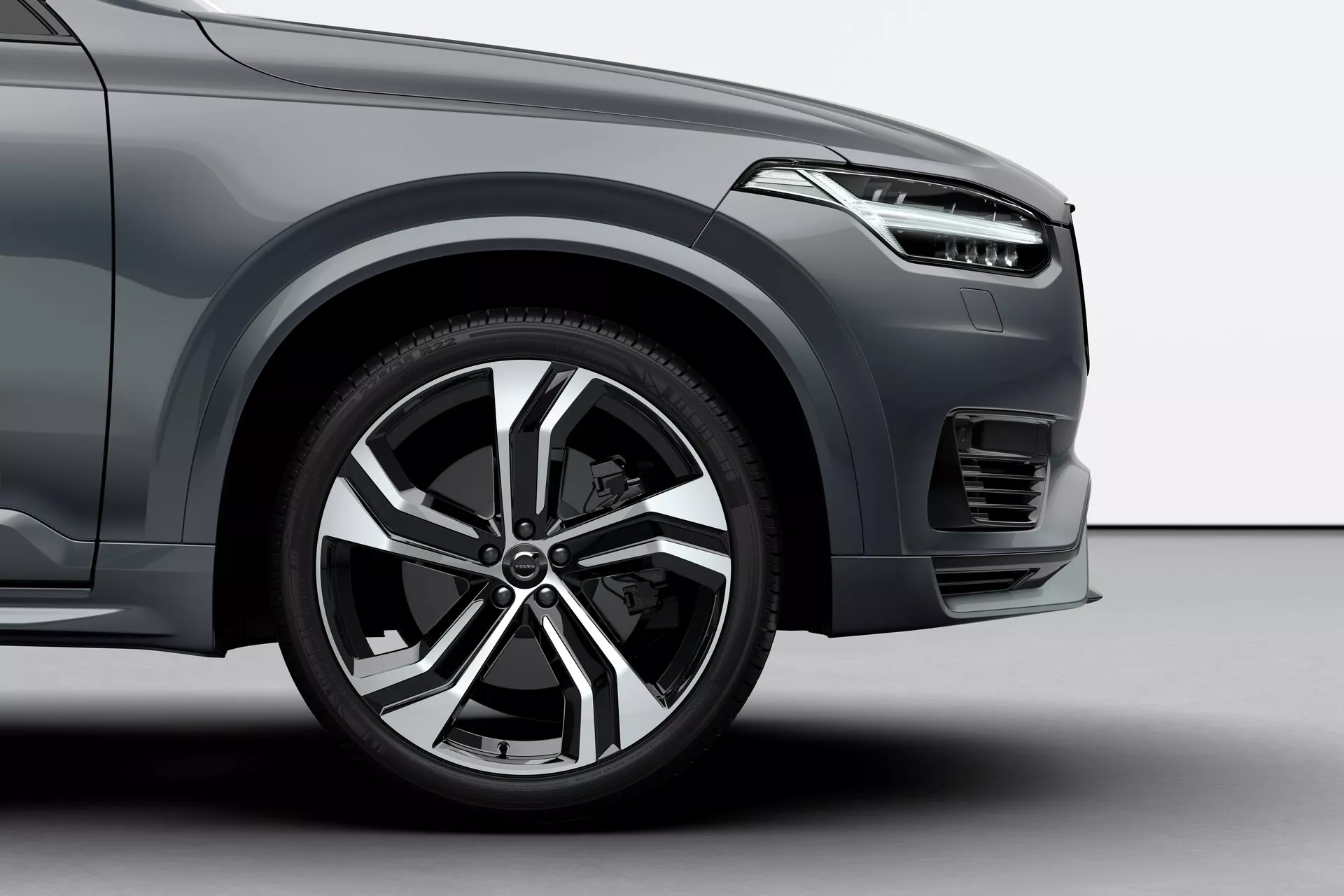 Toma en ángulo bajo delantera del lado del conductor de una Volvo XC90 2015, destacando el diseño moderno de la llanta de aleación con múltiples radios, el neumático deportivo de perfil bajo, parte del guardabarros delantero con líneas aerodinámicas, un diseño elegante del faro, el parachoques delantero con patrón de rejilla horizontal, una posible luz antiniebla o ventilación debajo del faro, y acabado de pintura metálica, todo lo cual sugiere un vehículo de lujo o de nivel superior.