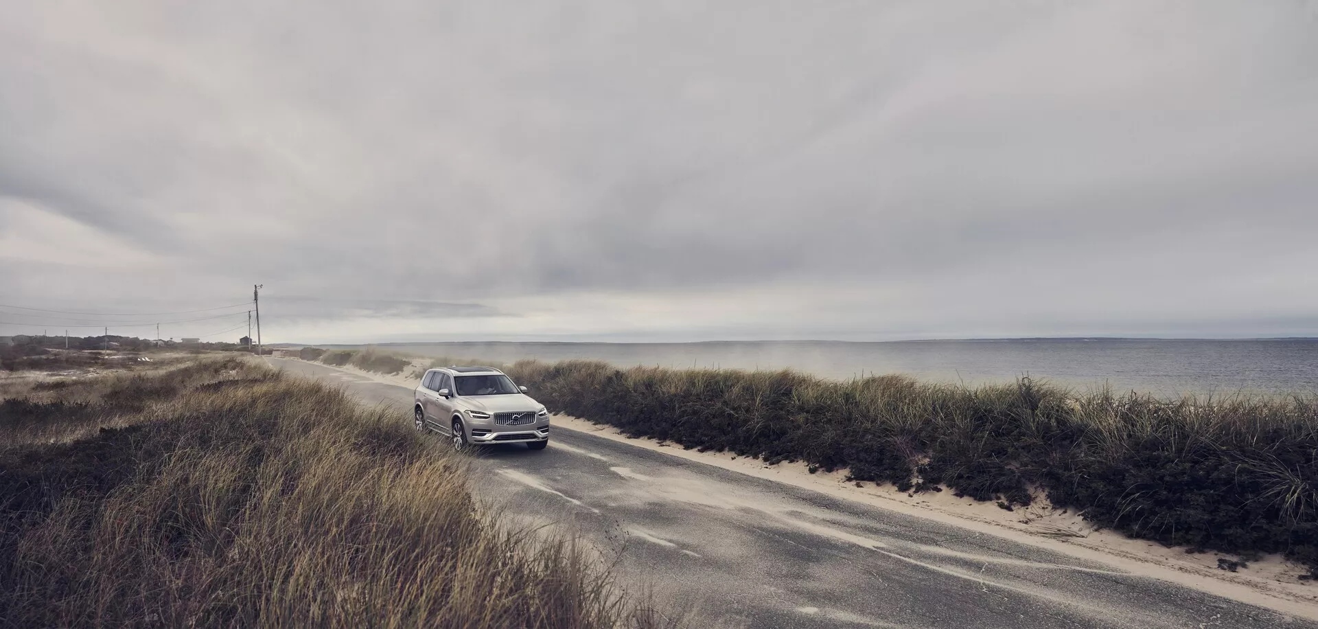 Foto de gran angular de un Volvo XC90 2015 (2da generación) conduciendo en una carretera costera estrecha. El coche se acerca a la cámara, destacando la parrilla frontal, faros y parachoques delantero. Con un cielo nublado y el océano en el fondo, el color metálico claro del SUV resalta. Esta escena enfatiza la combinación de lujo y aventura del vehículo, sugiriendo características tecnológicas y de seguridad avanzadas para las condiciones de conducción costeras.