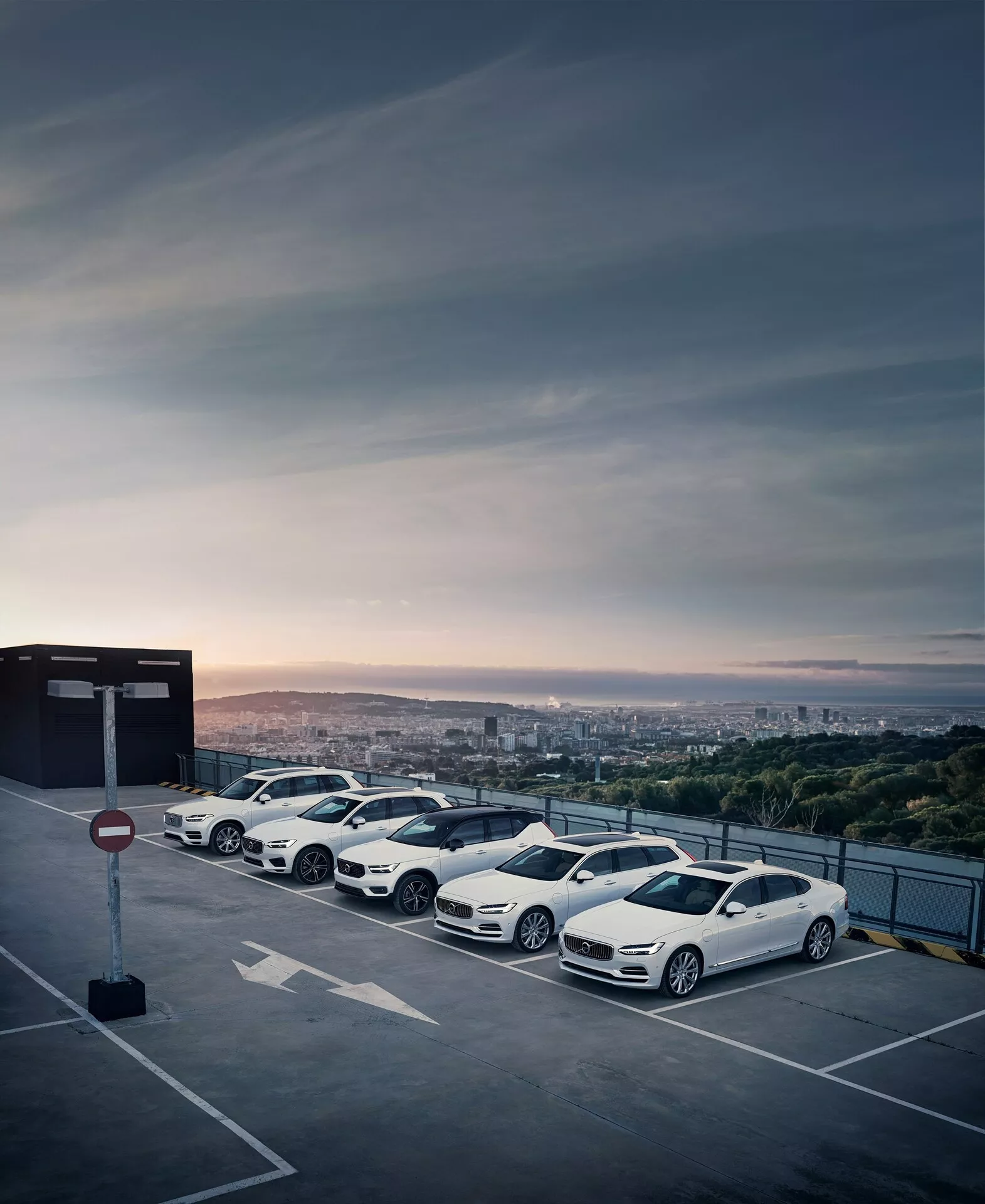 Línea de cuatro autos modernos y lujosos Volvo XC90 2015 (2ª generación) estacionados en el último piso de una estructura de estacionamiento elevada durante el amanecer o atardecer con un paisaje urbano de fondo. Los sedanes blancos presentan diseños aerodinámicos, llantas de aleación de varios radios, espejos retrovisores aerodinámicos y ventanas tintadas, mostrando sus contornos elegantes y estética dinámica.