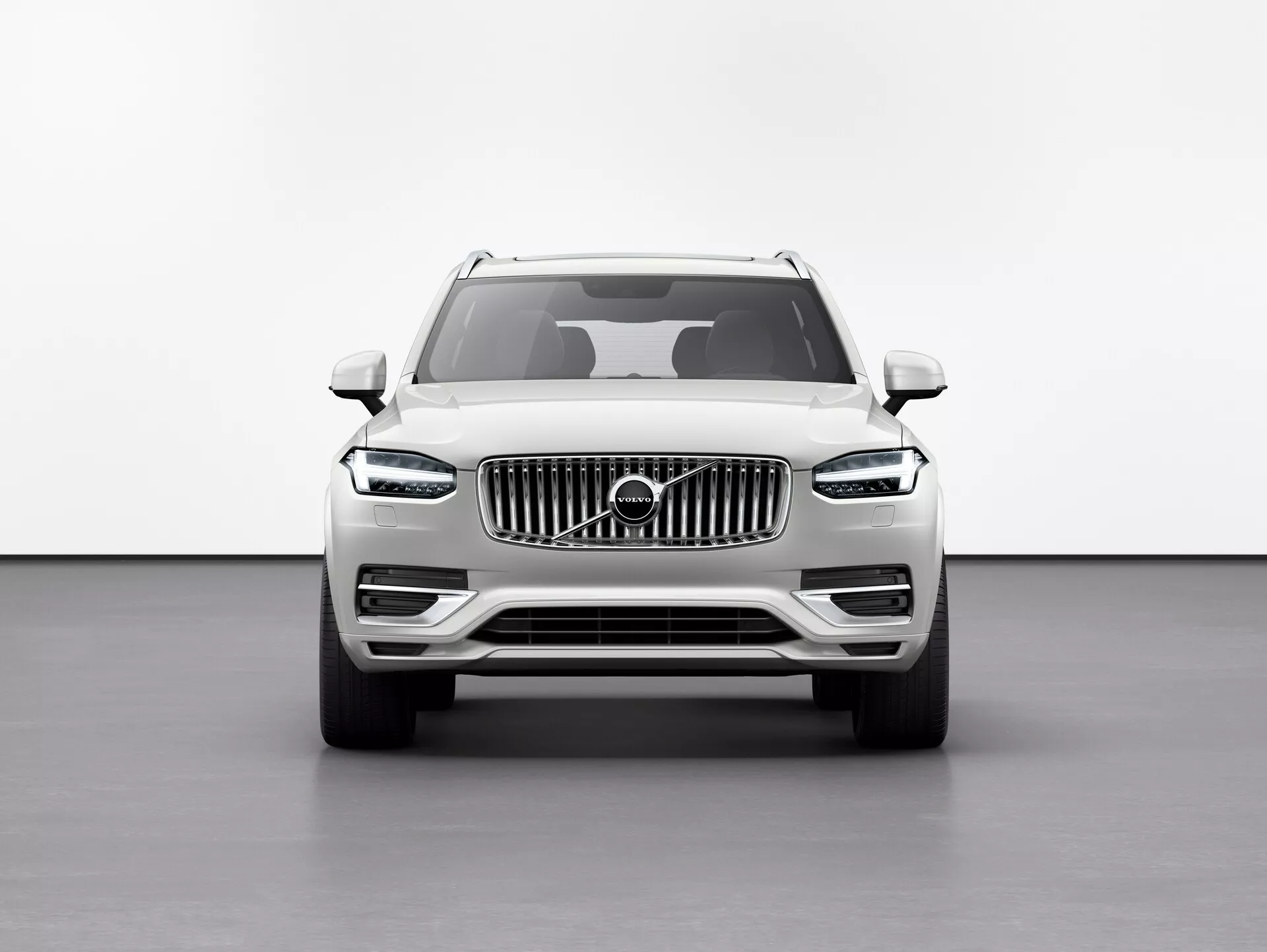 Vista frontal del Volvo XC90 2015 (2ª generación) SUV contra un fondo blanco en un entorno de estudio, destacando la altura elevada del vehículo, gran parrilla con barras verticales y emblema de Volvo, elegantes faros LED 'martillo de Thor', líneas definidas del capó, luces antiniebla en el parachoques inferior y espejos laterales con intermitentes integrados.
