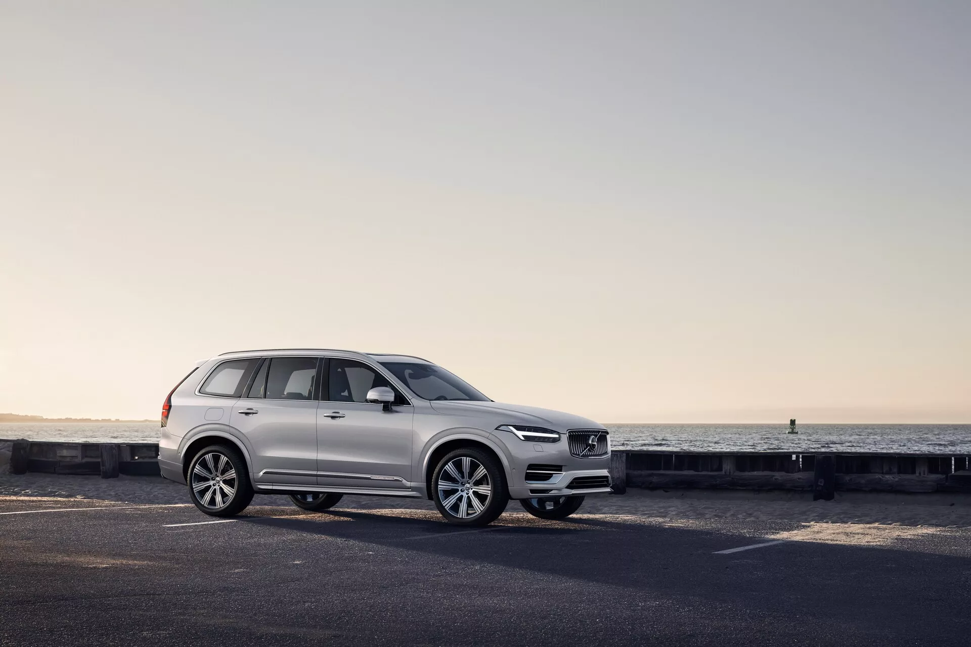 Vista de perfil lateral de un Volvo XC90 2015 SUV blanco en una carretera costera, con características de diseño visibles que incluyen el diseño de las ruedas, paneles de puertas, espejos laterales, rieles del techo y configuración de ventanas, acentuadas por la suave iluminación del amanecer o anochecer.