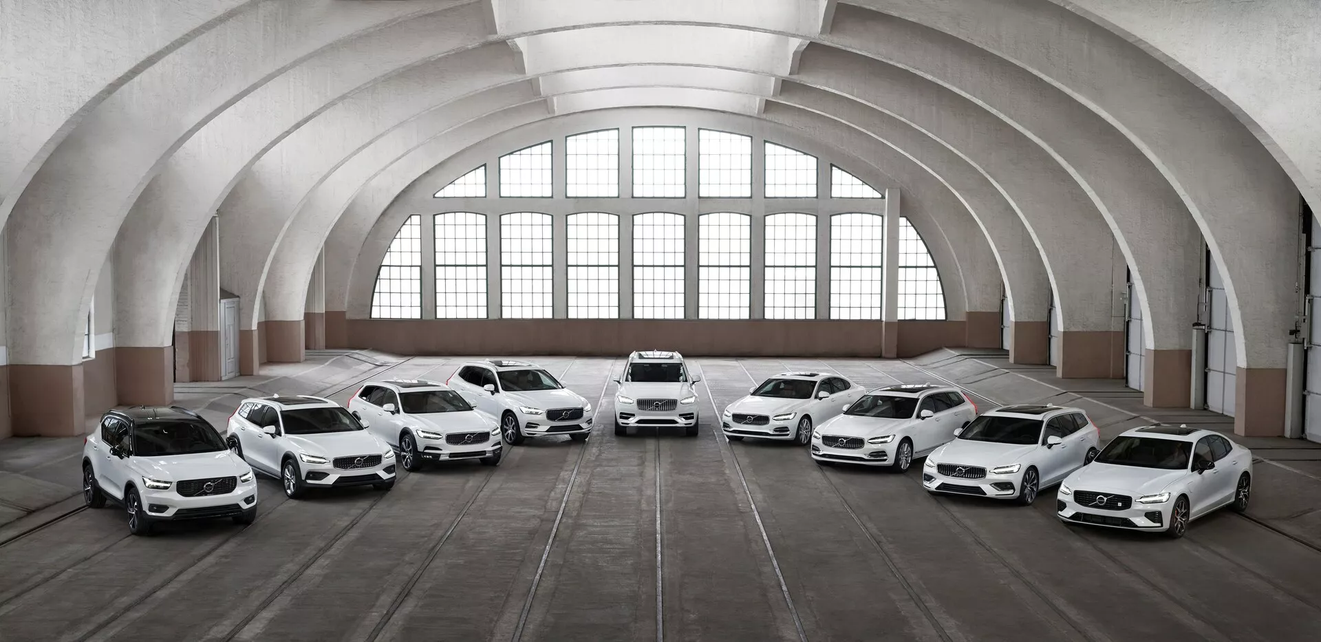 Una fila de ocho coches Volvo XC90 2015 de segunda generación, blancos, posicionados dentro de un espacioso edificio arquitectónicamente clásico con grandes ventanas arqueadas y un techo curvo. Los vehículos están orientados hacia la cámara en diferentes ángulos, mostrando sus frentes y características de diseño, incluyendo faros LED, grandes ruedas de aleación y paneles de carrocería esculpida. La iluminación resalta los elementos de diseño de los coches mientras que el suelo, marcado por huellas de neumáticos, sugiere el uso del espacio para alojamiento de vehículos.
