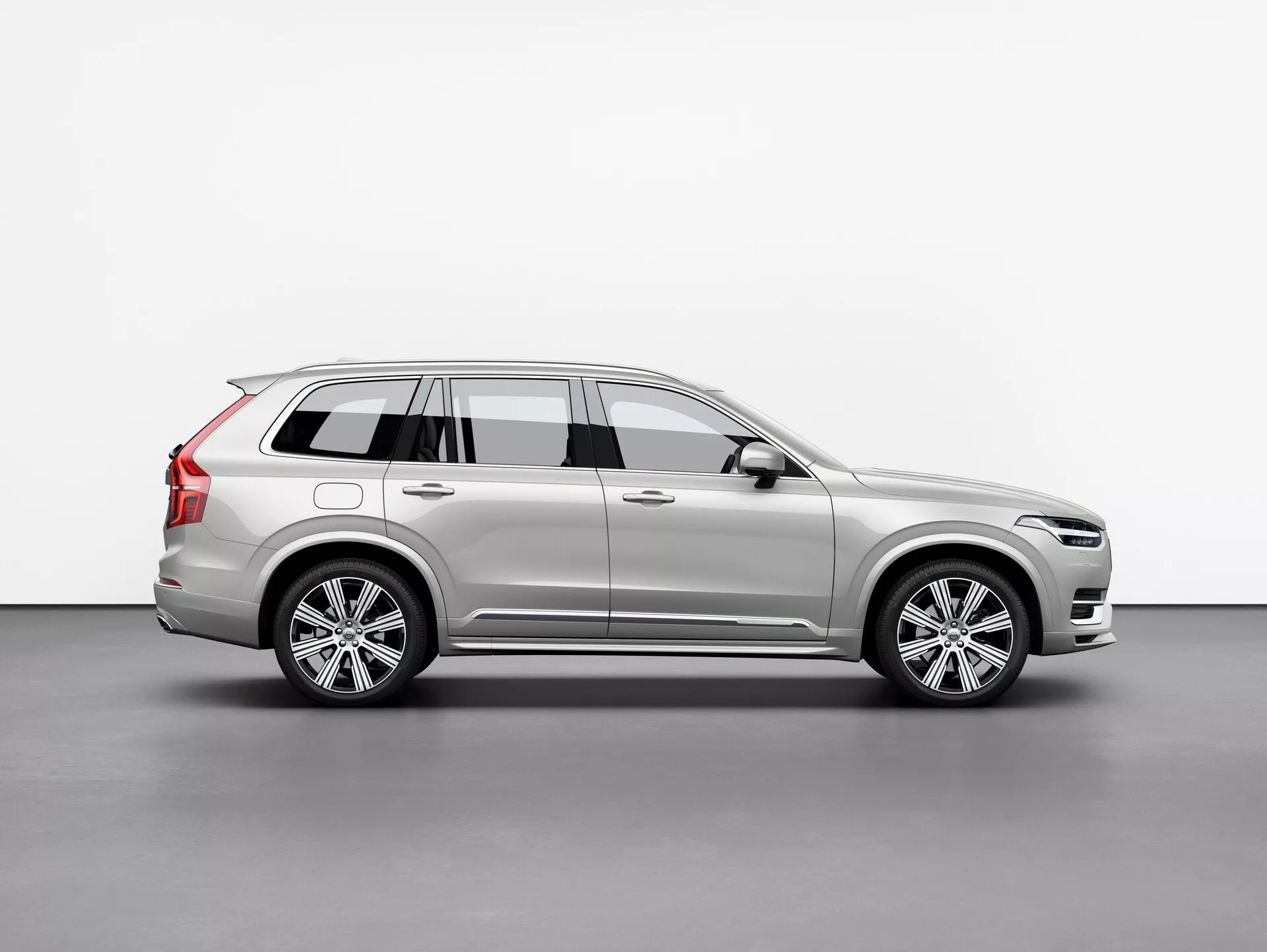 Imagen de perfil lateral de un Volvo XC90 2015 (2ª generación) SUV plateado, mostrando sus llantas de aleación, rieles de techo, espejo lateral con señal de giro integrada y líneas de carrocería detalladas, posicionada centralmente contra un fondo simple.