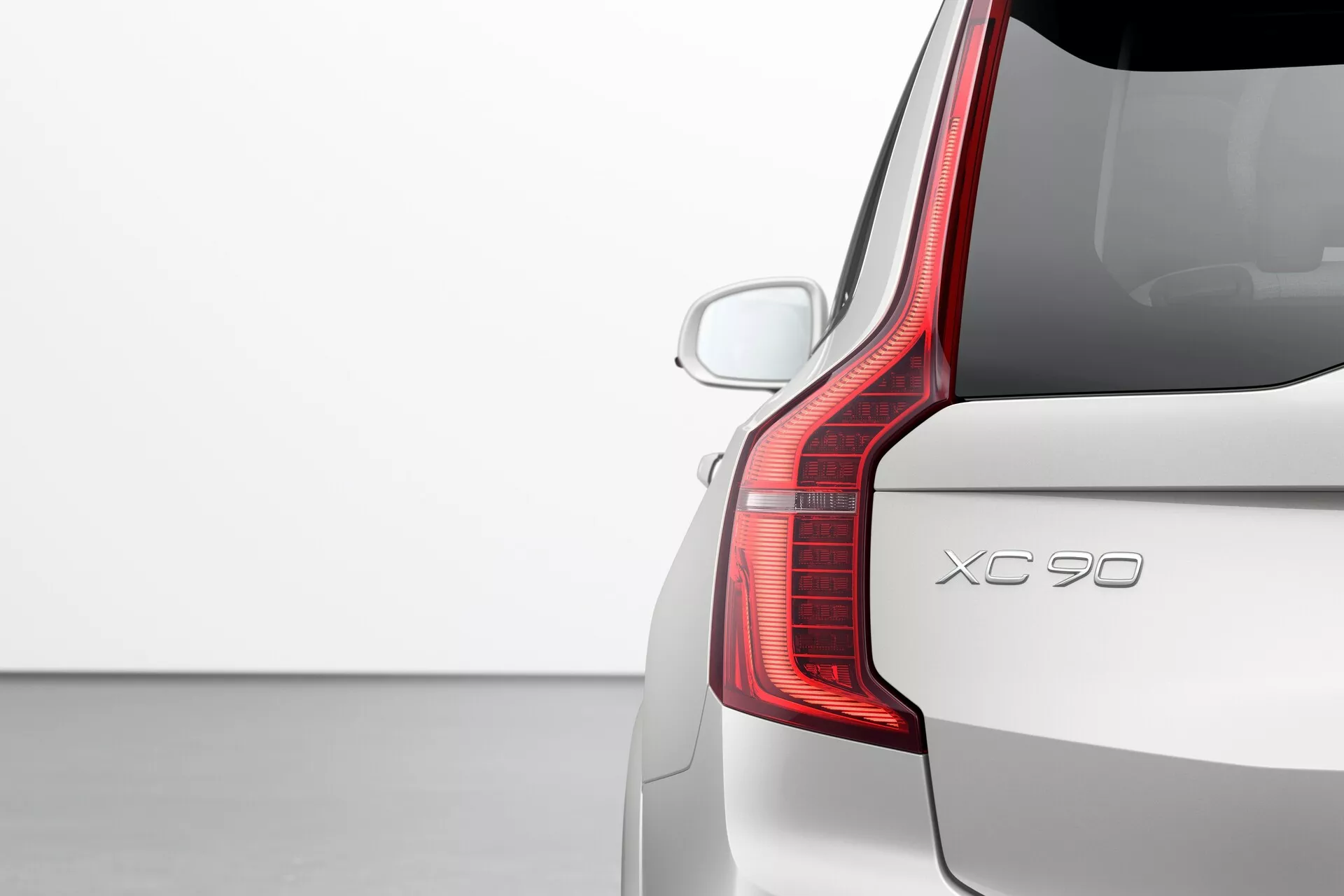 Vista detallada del cuarto trasero en el lado del pasajero de un Volvo XC90 2015 (2ª generación), mostrando el distintivo diseño de las luces traseras LED, la tapa del maletero y el parachoques trasero. El ángulo trasero de tres cuartos resalta la extensión vertical de la luz trasera y su forma única, que se curva hacia adelante en el lado. La imagen también destaca el parabrisas trasero con el alerón de techo aerodinámico, un sensor de estacionamiento trasero visible y la pintura de color sólido en un entorno de estudio simple, centrado en el diseño y características del coche.
