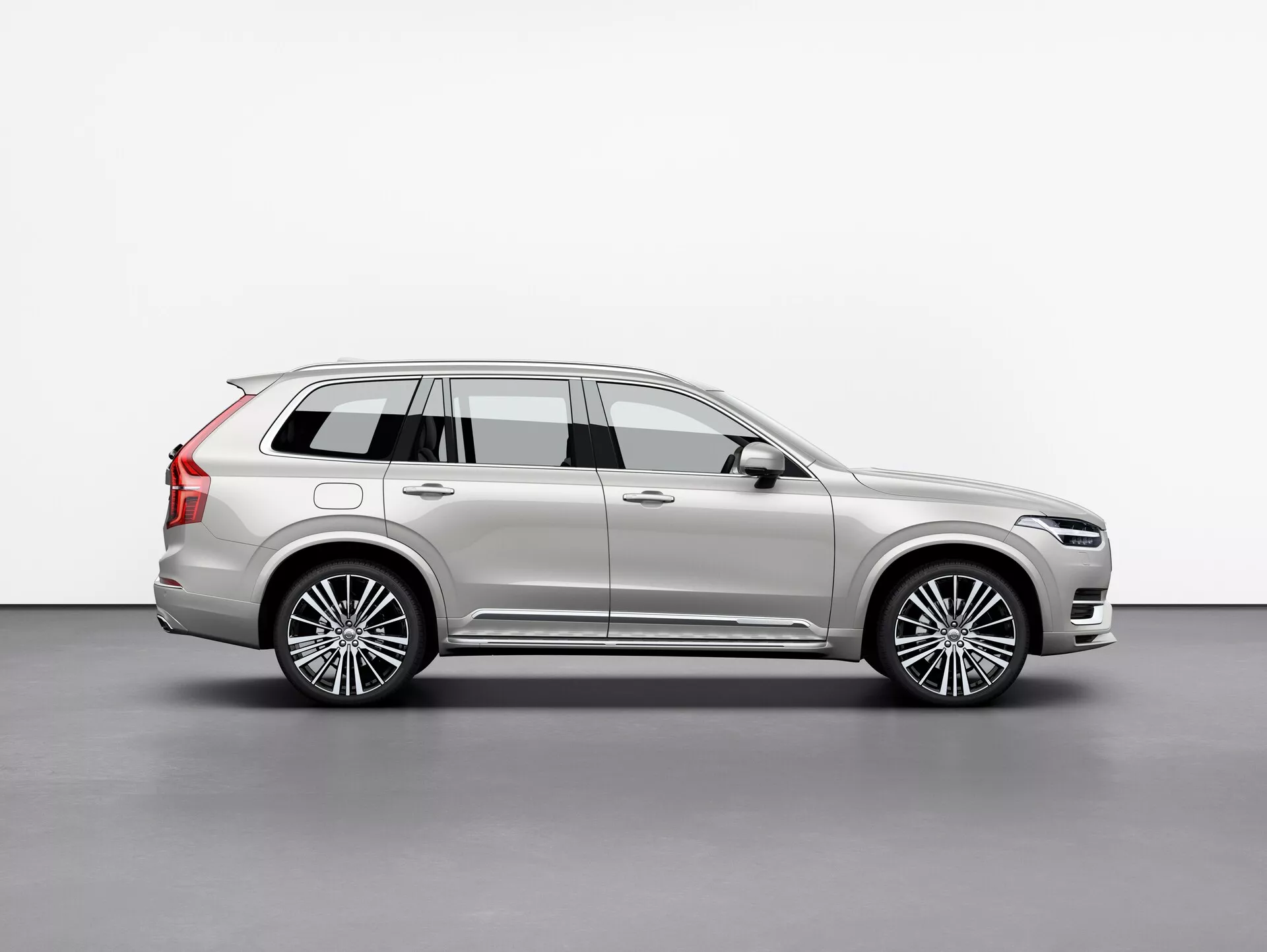 Perfil lateral del Volvo XC90 2015, un SUV de lujo de tamaño completo, capturado en un estudio con fondo blanco plano. La imagen muestra el largo entre ejes del vehículo, las cuatro puertas, el portón trasero, las llantas de aleación multirradio, los detalles en cromo, la pintura metálica y los elementos de diseño destacados como los espejos laterales y las barras del techo. La escena meticulosamente iluminada resalta las líneas y curvas de la carrocería, reflejando su naturaleza premium. El vehículo está posicionado para proporcionar una vista clara de su longitud, altura y postura general, indicativo de un SUV con capacidad para tres filas de pasajeros y carga.