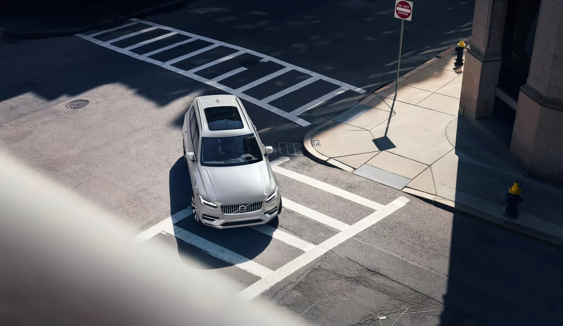 Vista aérea cenital de un coche Volvo XC90 plateado del 2015 situado en una intersección urbana durante el día. El vehículo, visto desde una alta perspectiva, muestra su techo panorámico, diseño elegante, rejilla, faros y parte superior de las puertas y espejos laterales. Los detalles de la calle incluyen líneas de paso de peatones, una línea de parada y señales de tráfico, con edificios altos y señales de calles indicativas de un entorno urbano. Las sombras sugieren que el sol está en un ángulo moderado, realzando la apariencia moderna y lujosa del coche. Características tecnológicas como sensores o cámaras son visibles en el parachoques delantero.
