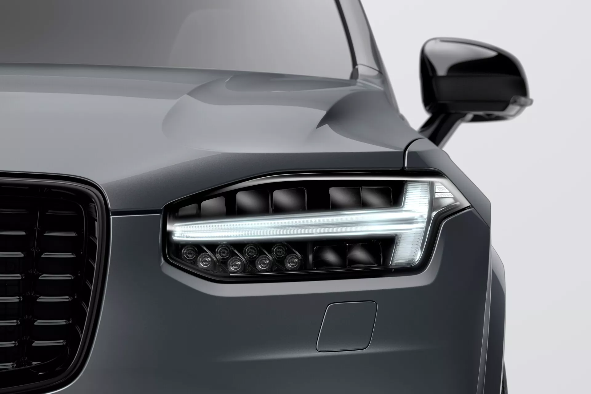 Primer plano del frontal de un Volvo XC90 2015 (2ª generación), destacando el faro izquierdo con tecnología moderna de LED o láser, elementos de iluminación dual, DRL elegantes y múltiples lentes de proyector para una mayor visibilidad. La imagen enfatiza la parrilla grande y estéticamente diseñada, el espejo lateral aerodinámico del color de la carrocería y el posible sensor ADAS debajo del faro. El acabado metálico gris oscuro suave y reflectante resalta las líneas elegantes pero musculosas del capó y el guardabarros, demostrando una pintura de alta calidad y un diseño de vehículo de vanguardia.
