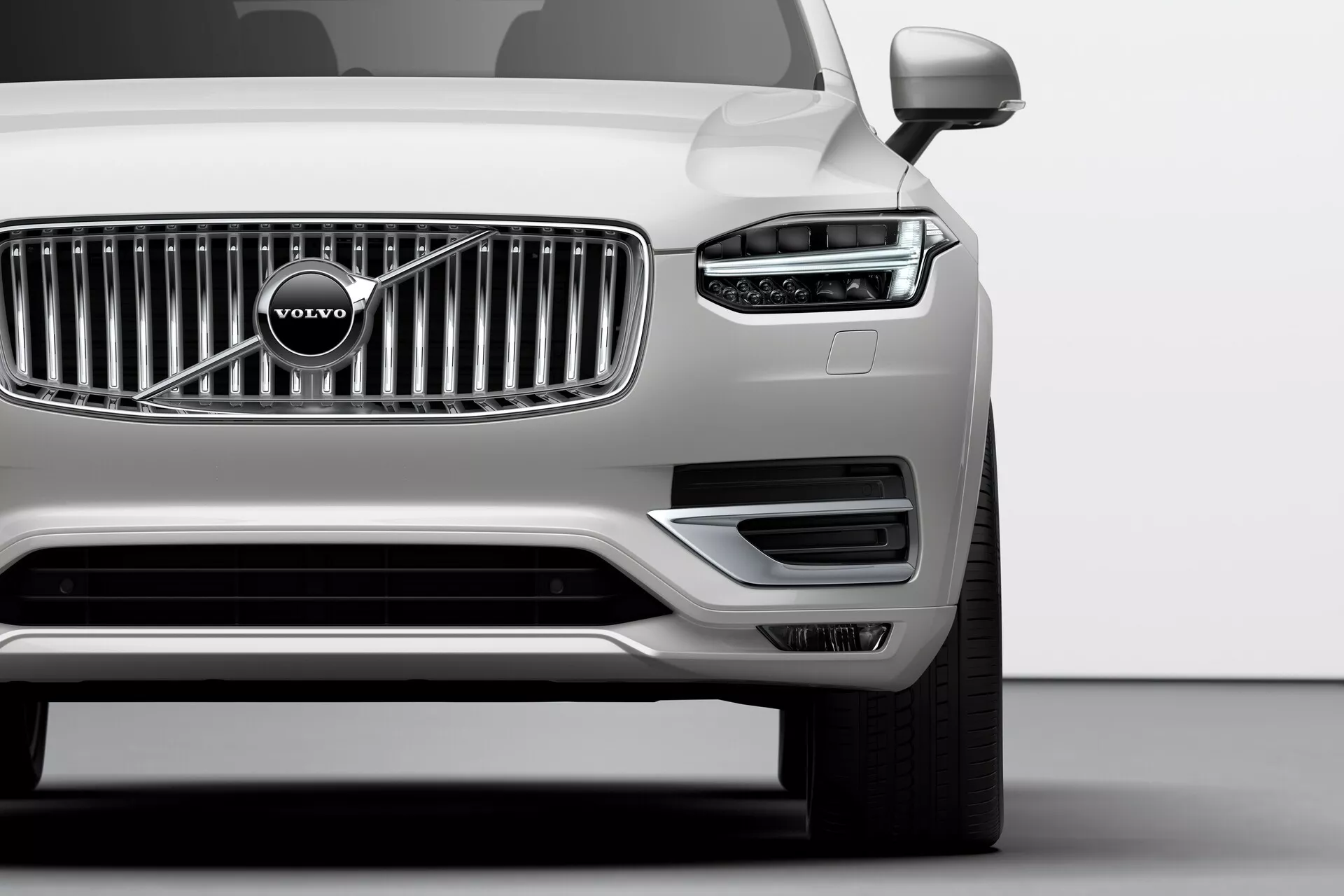 Toma en ángulo bajo del cuarto delantero izquierdo de un Volvo XC90 plateado de 2015 (2ª generación) en un estudio, mostrando la distintiva luz de conducción diurna LED en forma de Martillo de Thor, la parrilla de rejillas cromadas verticales con el emblema de Volvo, el parachoques delantero minimalista con entradas de aire, el espejo lateral izquierdo, la rueda de aleación multirradio y el impecable acabado metálico, ilustrando la tecnología avanzada de iluminación, el estilo de la marca y las características aerodinámicas.