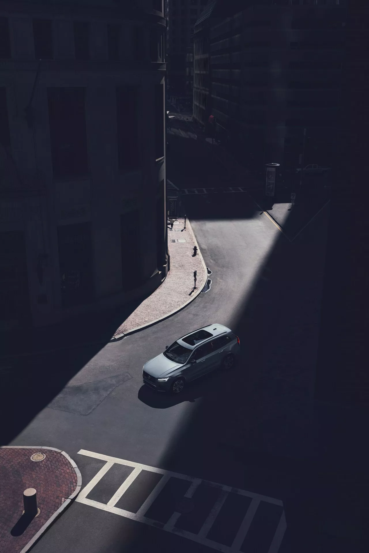 Vista aérea del sedán Volvo XC90 2015 (2ª generación) estacionado en una calle urbana sombreada, capturada desde un punto elevado mirando hacia abajo en un ángulo oblicuo. El maletero, el techo y el parabrisas trasero del coche son claramente visibles, con la parte trasera del vehículo iluminada por la luz del sol. La imagen captura la tapa del maletero trasero, la forma de la ventana trasera y la parte superior del maletero, mientras que las sombras ocultan los detalles específicos del diseño, como las luces traseras y el parachoques. La línea de techo aerodinámica es evidente, sin portaequipajes visibles ni techos solares. El coche está estacionado junto a un bordillo cerca de las fachadas de edificios y un cruce peatonal, enfatizando la soledad del vehículo en el entorno urbano sombreado.