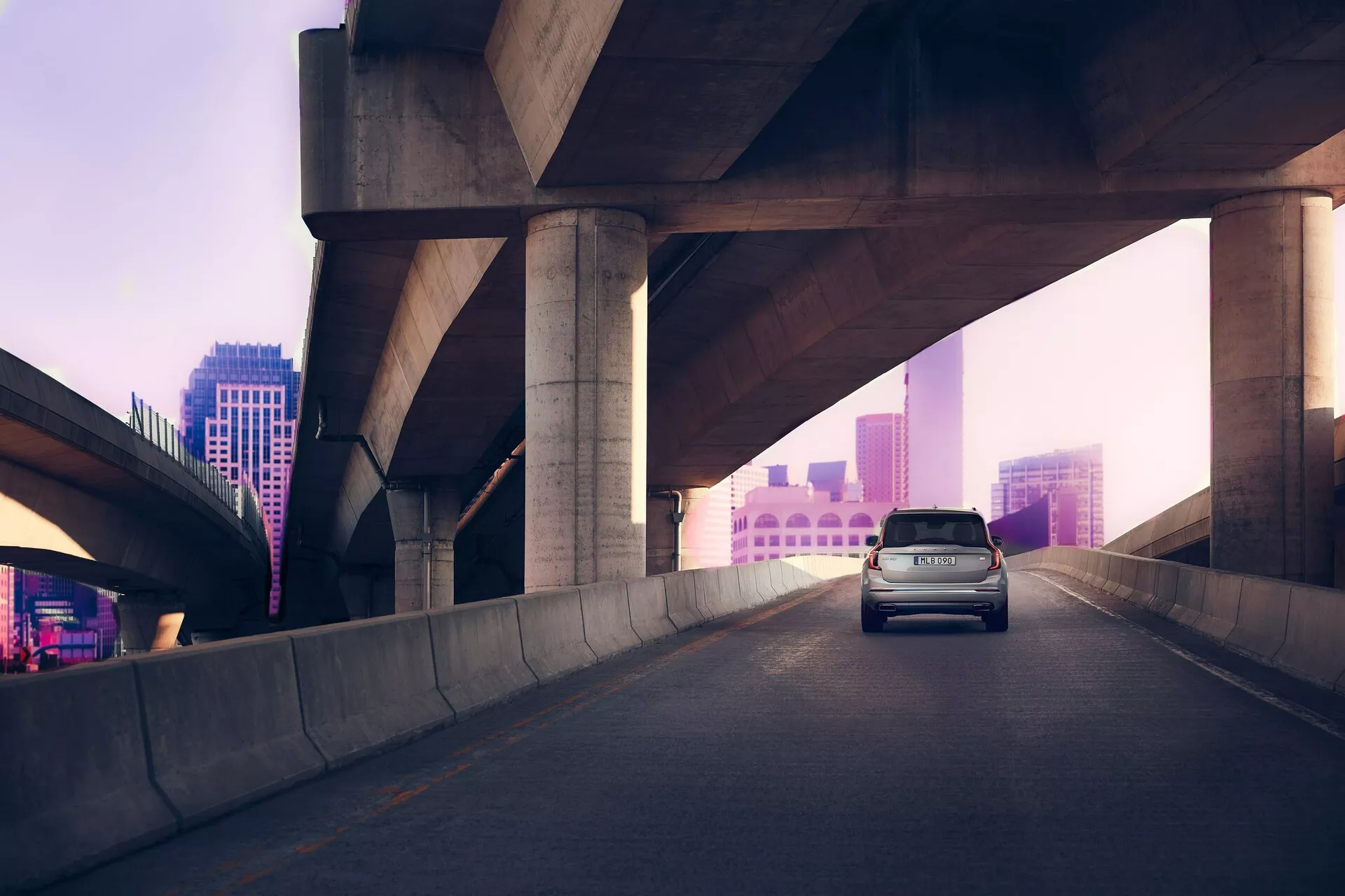 Vista trasera tres cuartos del Volvo XC90 de segunda generación 2015 en una carretera elevada, con un paisaje urbano suavemente iluminado en tonos rosa-púrpura al amanecer o anochecer. Las luces de freno del coche están encendidas, sugiriendo una desaceleración. El SUV cuenta con un techo alto, trasera tipo hatchback y diseño aerodinámico para eficiencia y estilo, capturado con luz natural difusa.