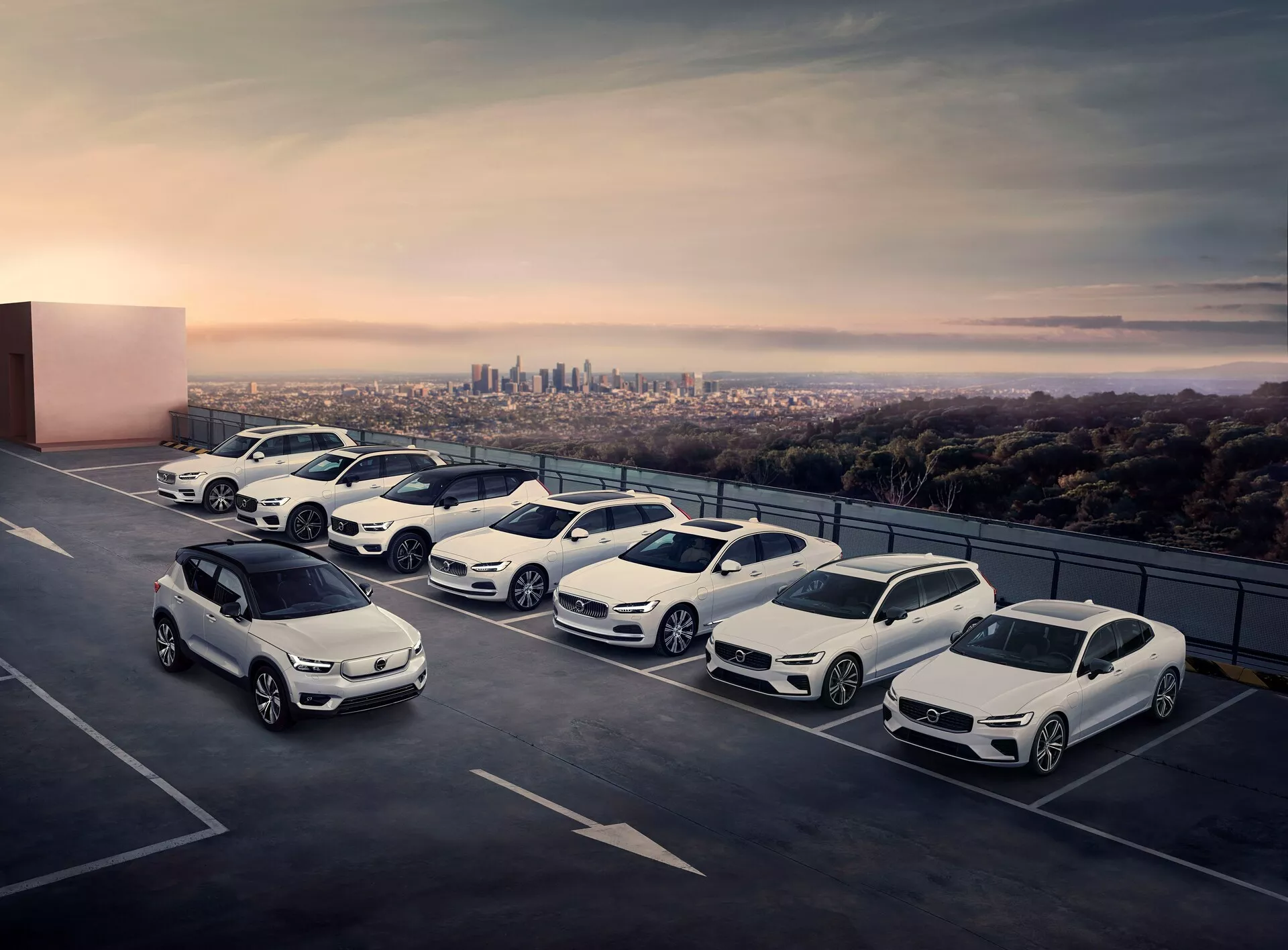 Vista elevada de cinco coches estacionados en diagonal en la planta superior de un aparcamiento de varios pisos, con el horizonte de la ciudad al fondo. Los vehículos incluyen una mezcla de SUVs compactos, sedanes y modelos más deportivos, todos con elementos de diseño moderno como faros LED y ruedas de aleación. La escena, capturada durante el crepúsculo, tiene una atmósfera serena y de alta gama.