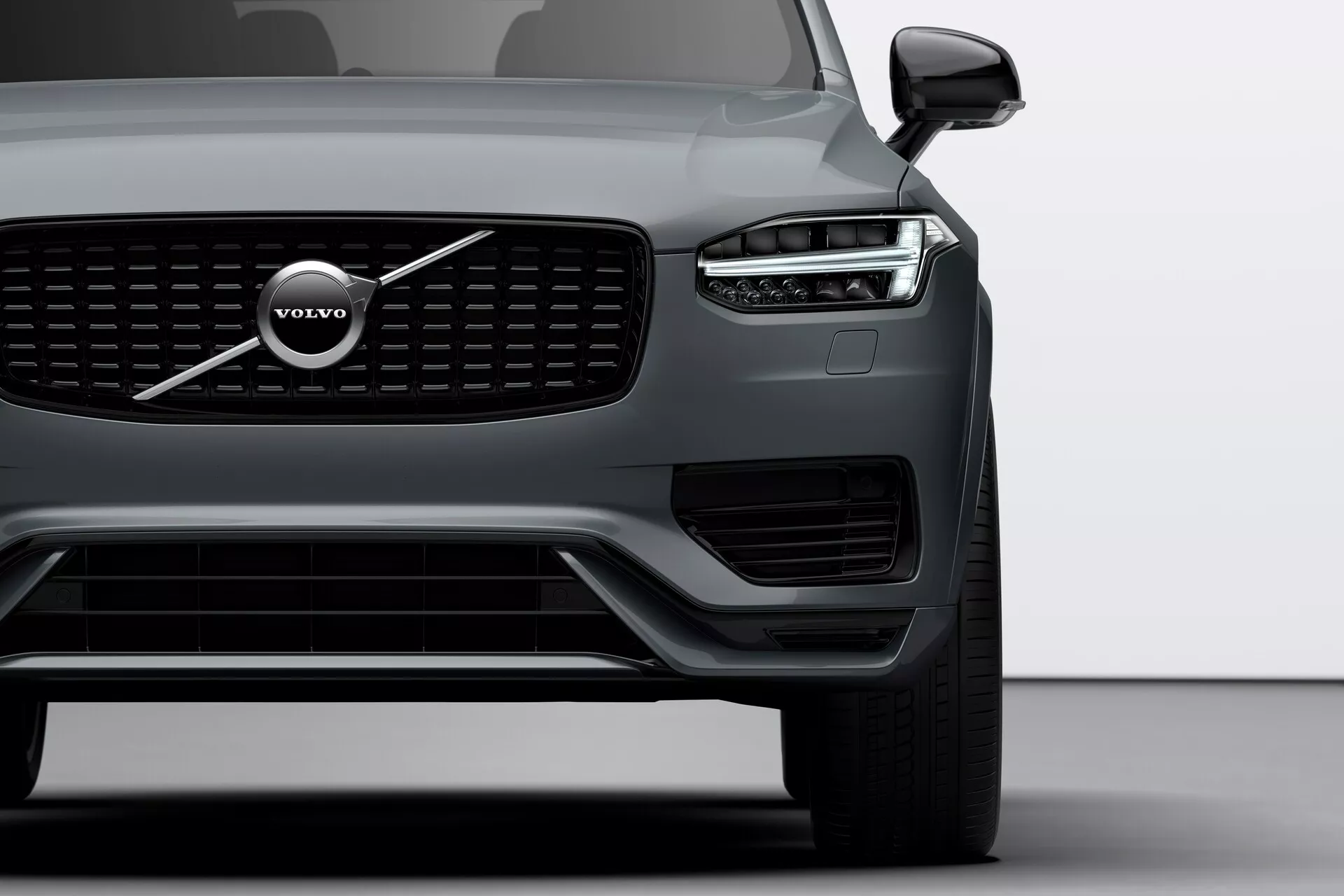 Vista en primer plano del ángulo frontal del Volvo XC90 2015 (2ª generación) centrado en su lateral izquierdo, destacando la distintiva parrilla, el gran emblema de Volvo y el diseño de faros LED 'Martillo de Thor'. La imagen incluye el faro, la rueda delantera, parte de la puerta del conductor, sensores visibles en el parachoques inferior y el espejo lateral con posible tecnología de monitoreo de punto ciego.