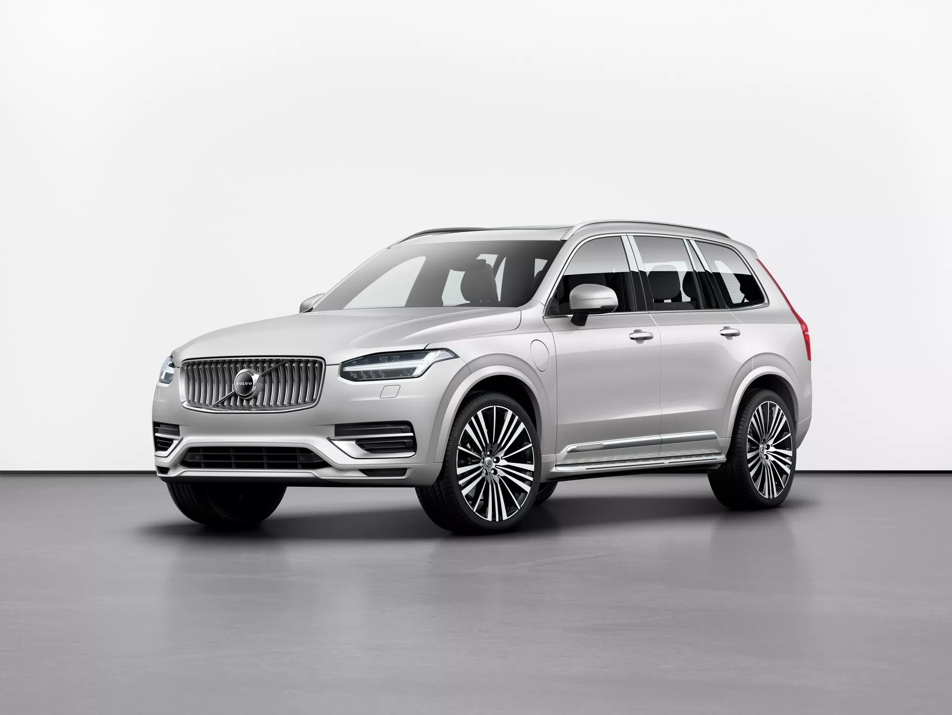 Fotografía de estudio de un SUV de lujo Volvo XC90 2015 (2ª generación), representado desde un ángulo frontal de tres cuartos, que muestra la rejilla frontal con un logotipo distintivo, faros LED elegantes, un parachoques delantero considerable y una larga distancia entre ejes con grandes llantas de aleación. Los espejos laterales se encuentran debajo del pilar A, con ventanas adornadas con molduras cromadas y barras de techo. El color de pintura neutro exuda sofisticación, y la imagen destaca las líneas y contornos del coche con una iluminación uniforme contra un fondo liso. Las características posibles incluyen tecnologías de asistencia al conductor y sistemas de información y entretenimiento avanzados.