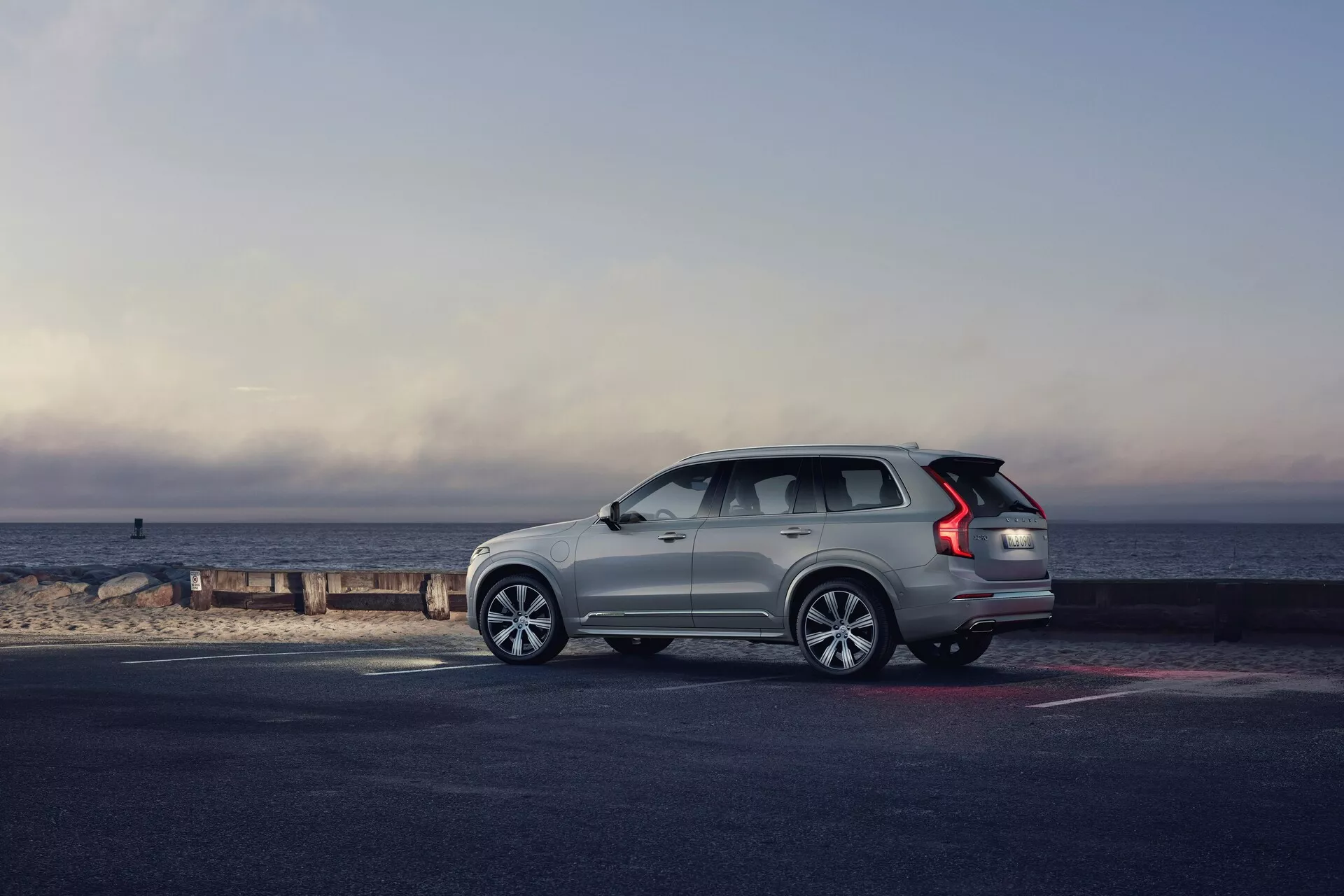 Vista de perfil de un SUV Volvo XC90 2015 (2ª generación) estacionado junto a la orilla, con el vehículo mirando hacia la derecha. El ángulo enfatiza el lateral y la parte trasera, mostrando elementos de diseño como las ruedas de aleación de varios radios, las luces traseras LED alargadas, las barras en el techo, la línea de las ventanas que asciende hacia la parte trasera y las manillas de las puertas al ras. El SUV de color claro destaca sobre el pavimento más oscuro y la playa, bañados por la luz natural de la mañana o última hora de la tarde, con un bonito fondo del horizonte donde el cielo se encuentra con el mar. La composición general evoca un sentido de aventura, lujo y libertad.
