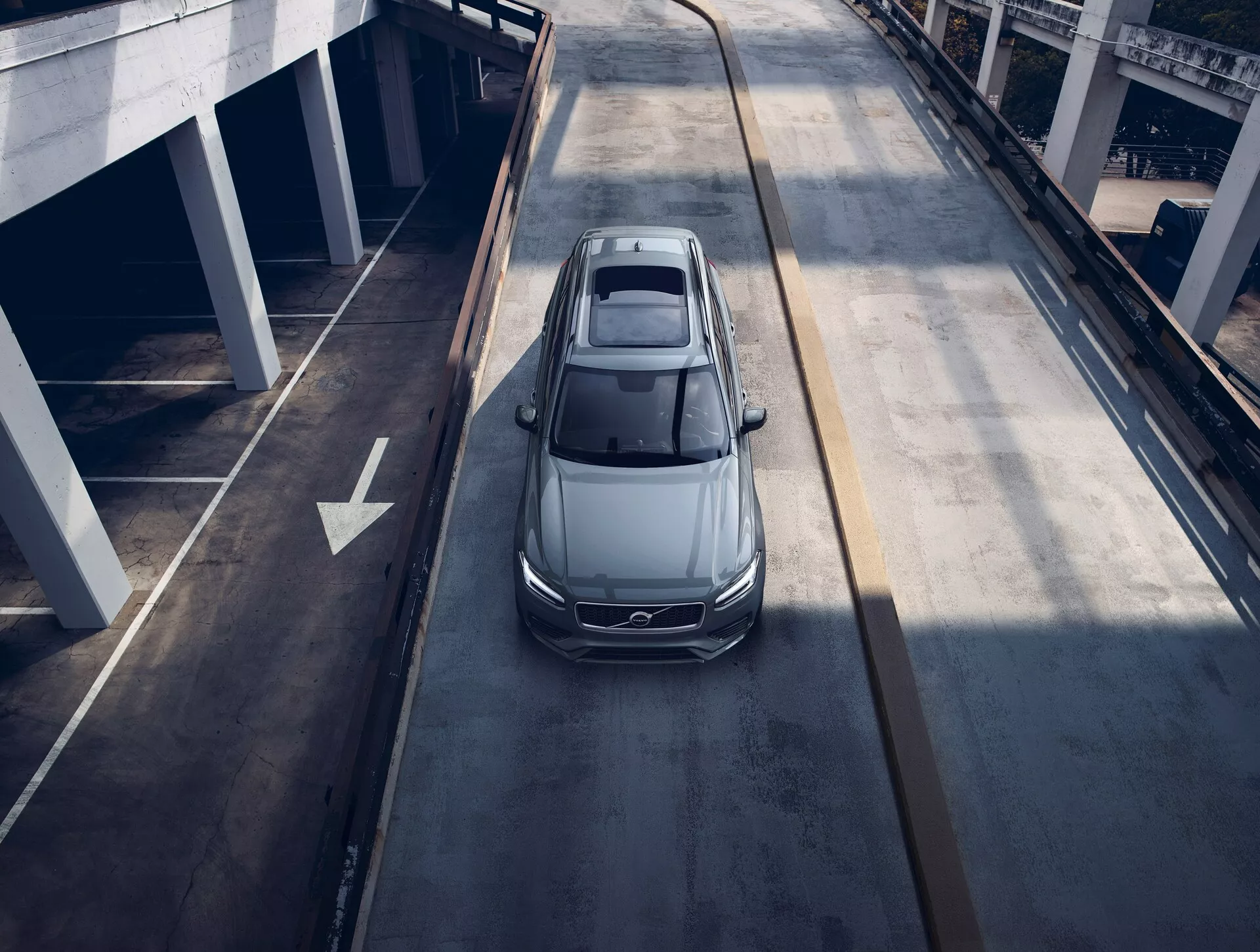 Vista de pájaro de un sedán Volvo XC90 2015 de color gris o plata, viajando por una carretera urbana de varios niveles, mostrando su techo solar panorámico, diseño aerodinámico, elegantes faros LED y grandes llantas de aleación, sugiriendo un vehículo moderno y lujoso.
