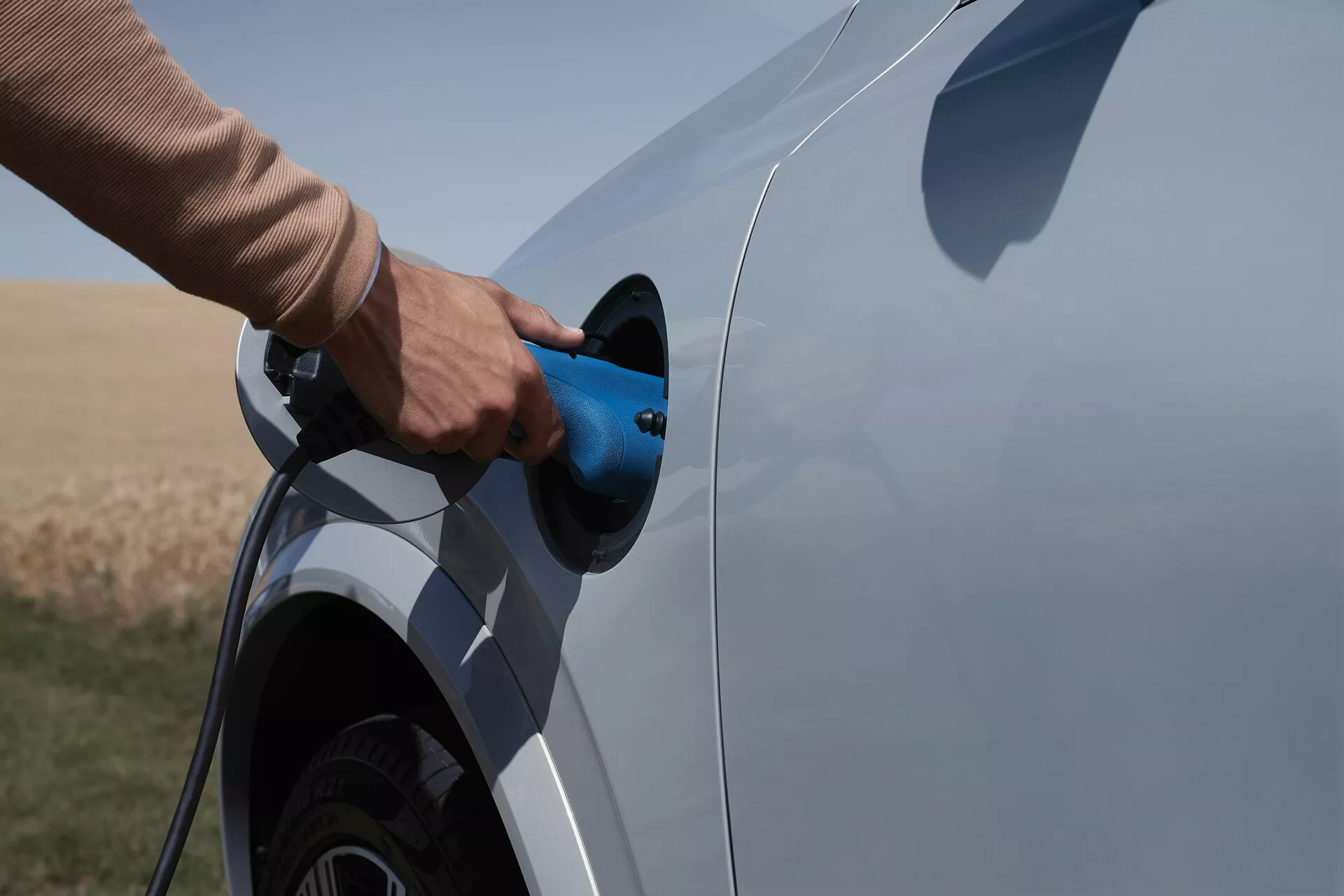 Vista lateral de un Volvo XC90 2015 de segunda generación eléctrico o híbrido enchufable durante el proceso de carga. La toma captura el lado del vehículo con enfoque en el puerto y el enchufe de carga, mostrando parcialmente la rueda trasera y una parte del fondo del coche. Cable de carga azul siendo insertado, fondo de área natural abierta, indicando un entorno rural.