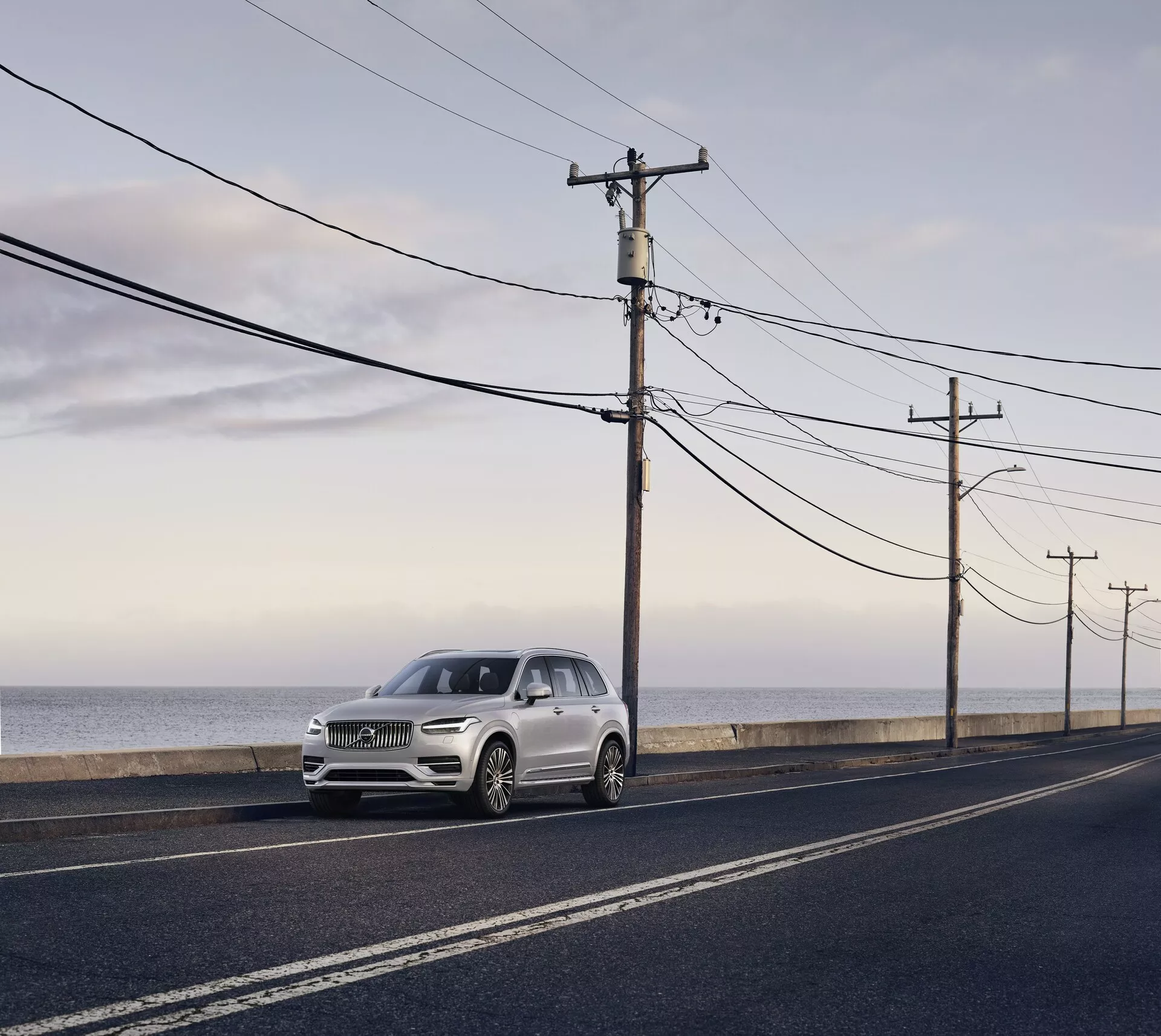 Volvo XC90 de segunda generación de 2015, SUV de lujo estacionado en una carretera de asfalto paralela a la costa del océano, capturado a la luz del día con luz difusa suave y sombras largas. Vista desde un ángulo lateral-frontal, destacando los elementos de diseño elegantes, incluidos los faros, la parrilla y los contornos del capó, color plateado claro o champán pálido que contrasta con la carretera oscura y el fondo natural.