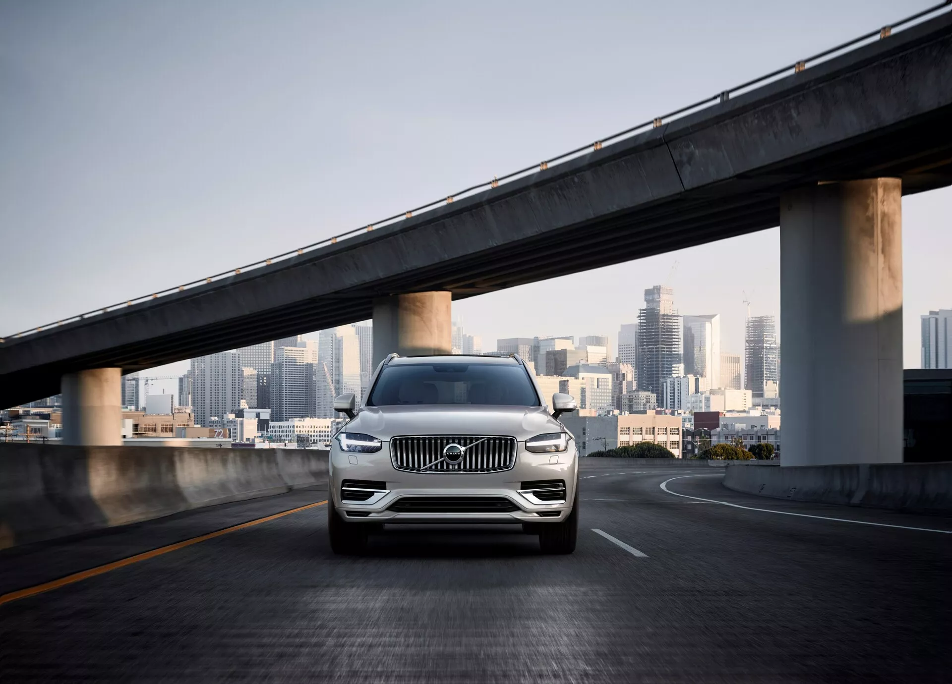 Una toma frontal del SUV Volvo XC90 de segunda generación de 2015 en un paso elevado vacío, con el horizonte de la ciudad en el fondo. El ángulo de la cámara es bajo, mostrando la parrilla frontal, las luces LED, los acentos cromados, las llantas de aleación y un parachoques esculpido con sensores, indicando asistente de estacionamiento o sistema de detección de colisión. La pintura perlada o metálica del coche brilla a la luz del día, destacando su lujo y diseño moderno en un entorno urbano.