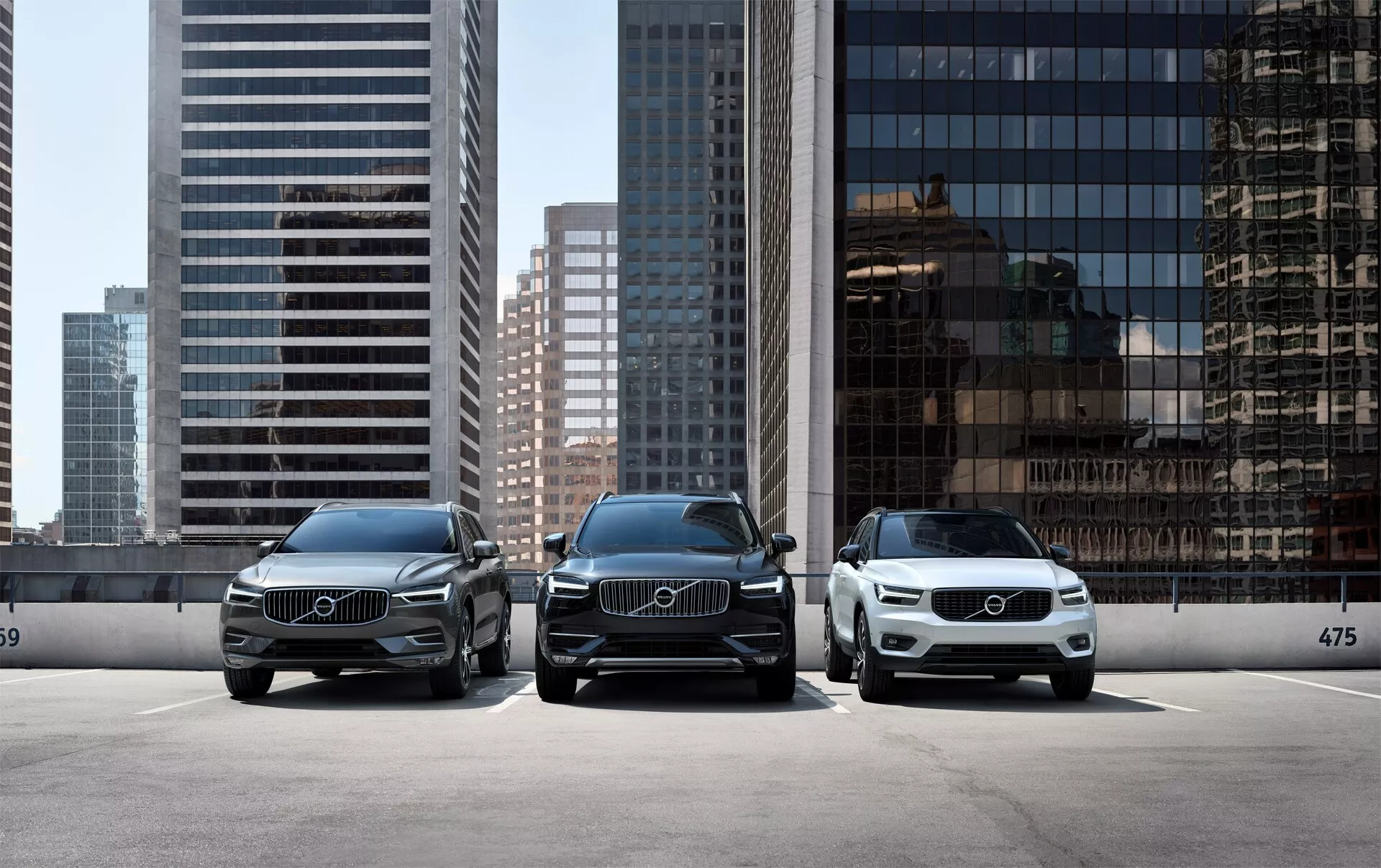 Tres autos de lujo Volvo XC90 2015 (2ª generación) estacionados en paralelo, de frente a la cámara, con un horizonte urbano moderno de fondo. La perspectiva frontal de los vehículos muestra faros LED elegantes, grandes parrillas con emblemas prominentes de Volvo y un diseño limpio y elegante. El auto en primer plano a la derecha es de un color más claro, destacando la variedad de opciones de color disponibles. Están estacionados detrás de un tope de borde con el número 475 visible, sugiriendo una zona de estacionamiento designada o una configuración de exhibición en una sala de exposición.