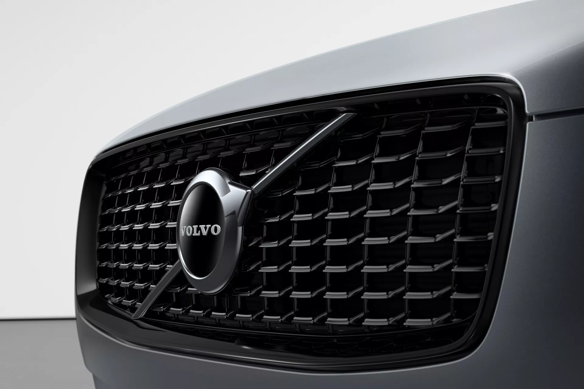Toma de primer plano de la sección frontal de un Volvo XC90 2015 (2ª generación) enfocada en la parrilla, con un patrón de malla densa con elementos rectangulares, un emblema de Volvo cromado en el centro con una flecha apuntando en diagonal hacia arriba a la derecha, y una vista del carrocería oscura y elegante del coche a la izquierda. La imagen resalta la estética del diseño y el reconocimiento de la marca.