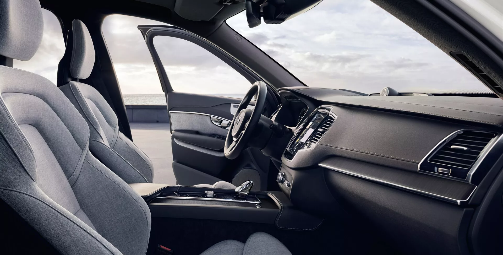 Esta imagen muestra el interior de un Volvo XC90 de segunda generación de 2015 desde la perspectiva del conductor. El ángulo ligeramente elevado ofrece una vista detallada de los asientos delanteros y el tablero, con un asiento del conductor a la izquierda tapizado en tela texturizada con un reposacabezas visible y soporte lateral. El volante multifuncional tiene varios botones de control y el salpicadero elegante incluye ventilaciones de aire simétricas y una pantalla de infoentretenimiento central parcialmente visible. La consola central muestra una palanca de cambios de transmisión automática y otras características de control, enfatizando un diseño ergonómico y práctico.