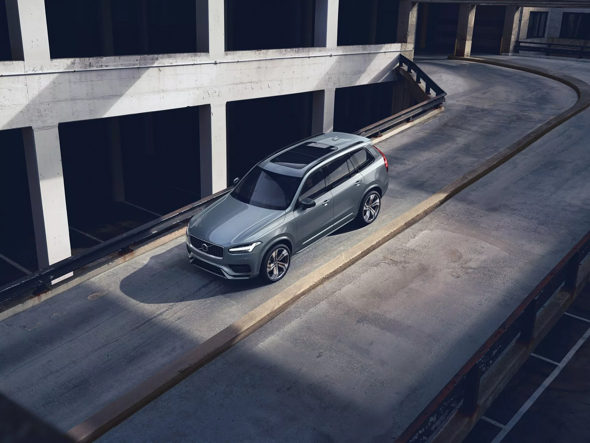 Vista de pájaro en ángulo alto de un SUV Volvo XC90 2015 estacionado en una superficie de concreto, mostrando su diseño moderno y elegante con acabado de pintura metálica, distintiva parrilla frontal, faros visibles y ruedas de aleación intrincadas. La toma destaca el techo, capó y lado izquierdo del coche, con la sombra proyectada que sugiere luz de la mañana o tarde. El entorno parece ser un nivel superior de una estructura de estacionamiento de varios pisos, realzando la prominencia del vehículo en un entorno urbano limpio.