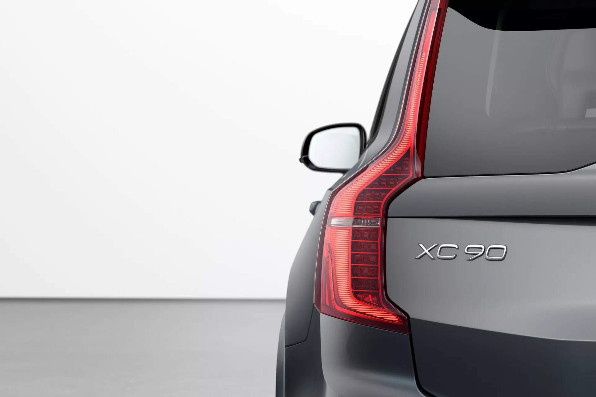 Vista de la esquina trasera de un Volvo XC90 2015 (2ª generación) resaltando detalles de diseño: luz trasera vertical LED, nombre del modelo 'XC90', parabrisas trasero con alerón, y acabado de pintura metálica premium sobre un fondo claro.