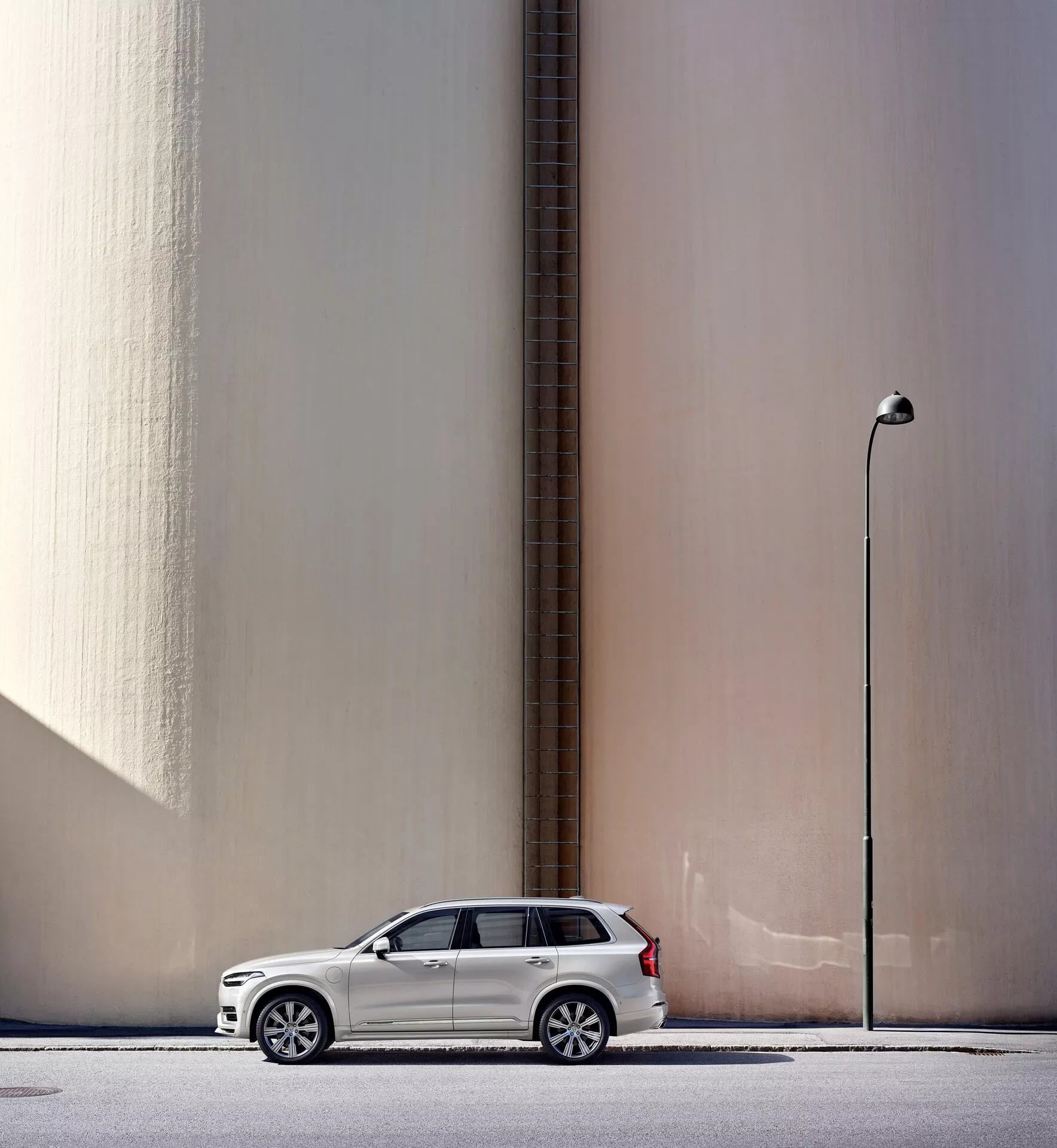 Vista de perfil lateral de un SUV Volvo XC90 2015 blanco estacionado en paralelo a una pared beige alta y lisa con una tubería vertical, con un telón de fondo urbano que incluye una lámpara de calle. El coche, posicionado a la izquierda del marco con los espejos laterales desplegados, proyecta una sombra ligera que indica luz natural del día. La imagen también destaca elementos de diseño moderno como ruedas de aleación y faros LED, subrayando características automotrices clave.