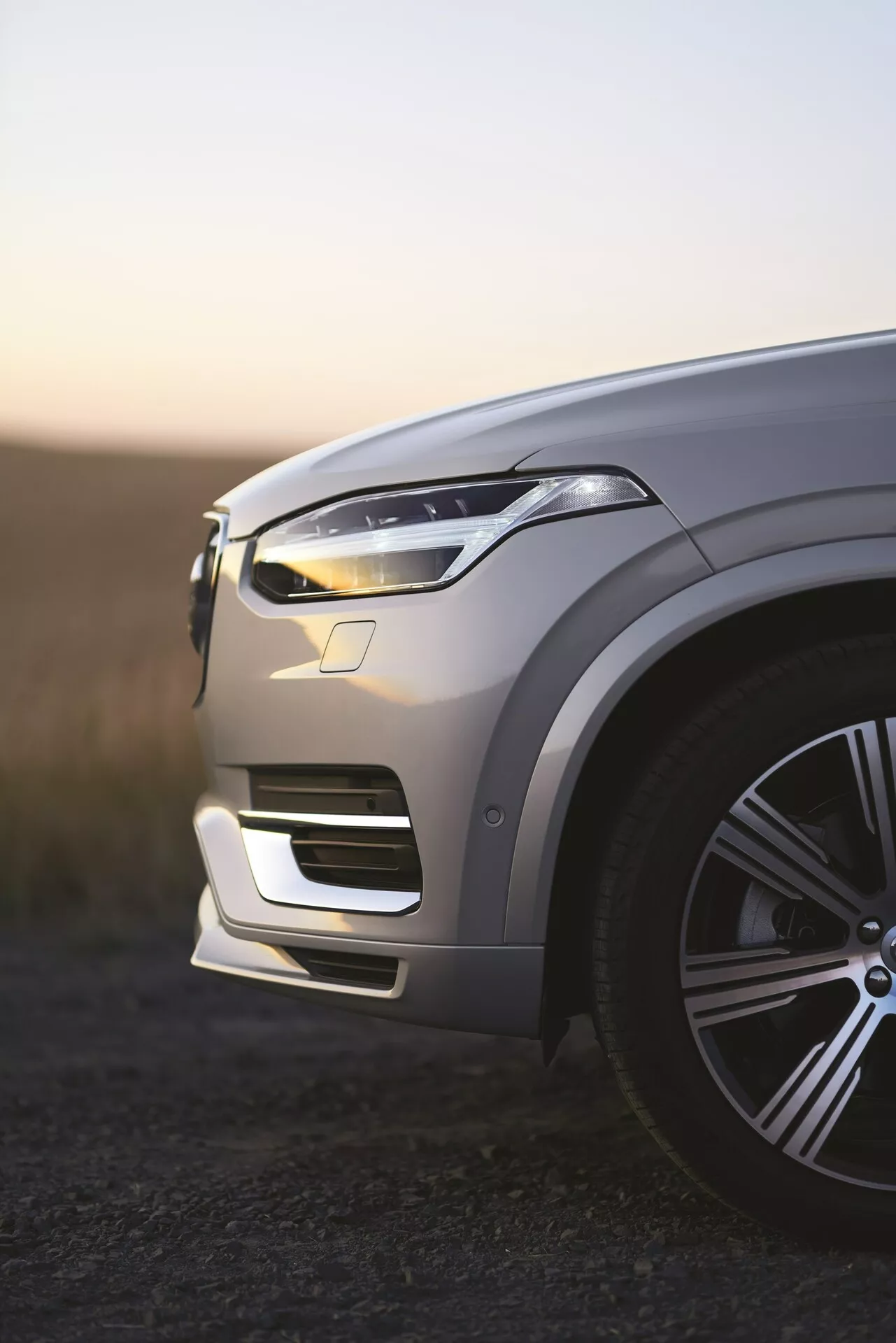 Vista del cuarto delantero izquierdo del SUV de lujo Volvo XC90 de segunda generación del 2015 al amanecer, luces diurnas LED iluminadas, pintura metálica que refleja la luz ambiental, elegante llanta de aleación grande, contornos intrincados del capó, diseño aerodinámico de luz LED, parachoques delantero integrado con tomas de aire, posibles sensores de asistencia al conductor visibles, profundidad de campo baja con fondo desenfocado.