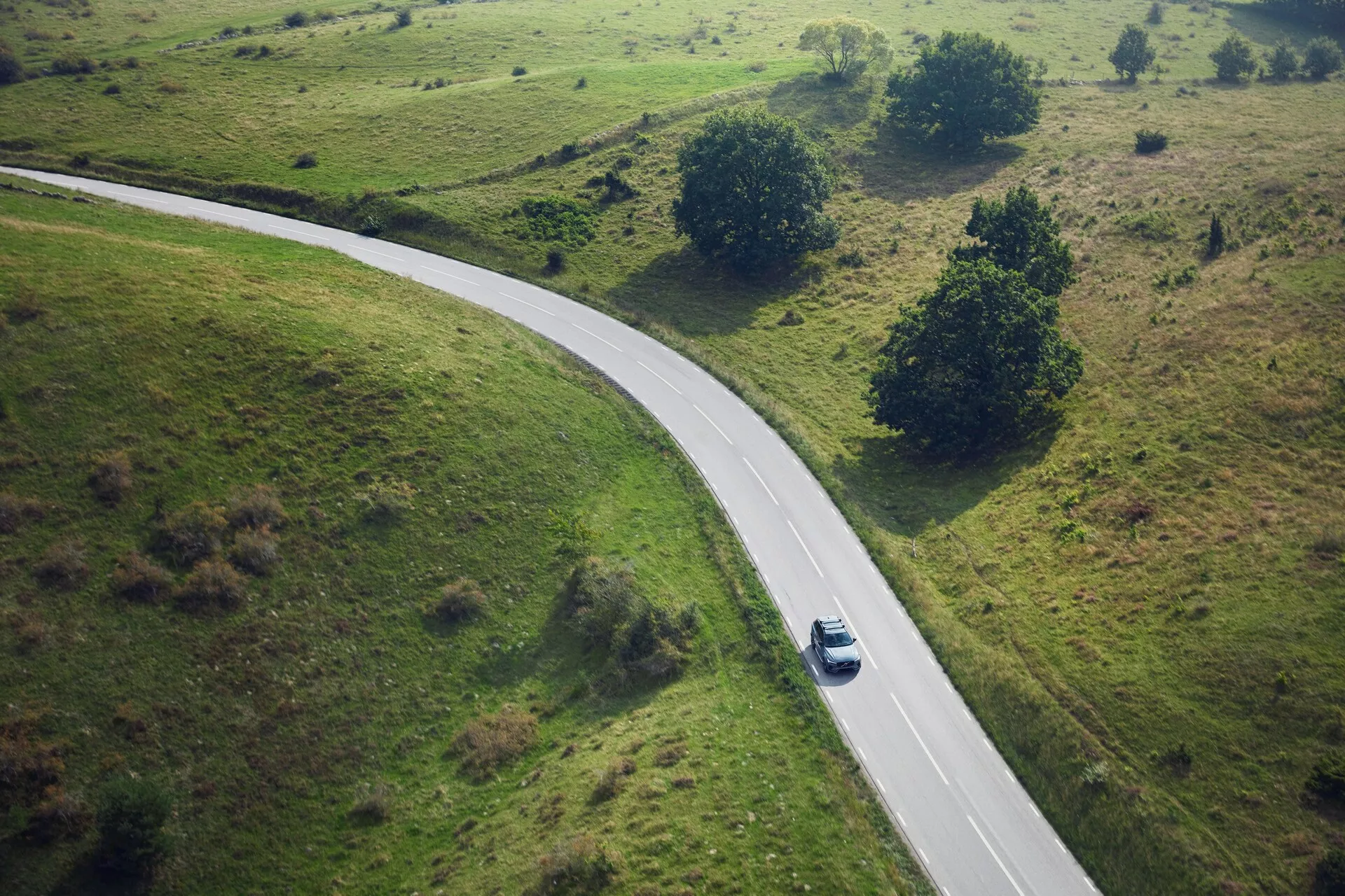 Vista aérea de un Volvo XC90 2015 plateado o gris claro conduciendo por un camino rural curvado rodeado de campos de hierba con pequeños árboles y arbustos. El coche, capturado desde una perspectiva de vista de pájaro, está posicionado a mitad de la curva en el centro derecho del encuadre, proyectando una sombra que sugiere sol de mediodía.