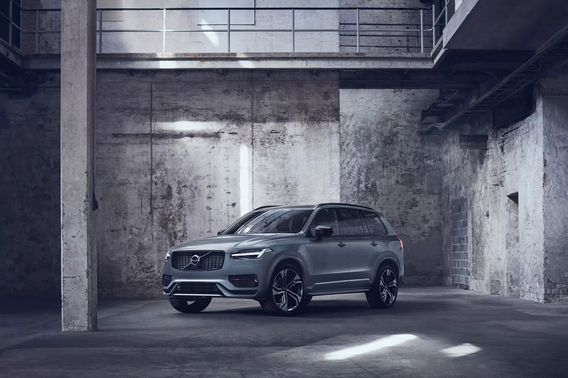 Imagen de un Volvo XC90 2015 de color metálico estacionado en un garaje de estilo industrial, débilmente iluminado. Capturado desde una vista frontal de tres cuartos, mostrando el lado del conductor con el perfil lateral y el frente prominentes. El coche cuenta con faros LED elegantes, una gran parrilla delantera, posiblemente sensores de estacionamiento y grandes llantas de aleación con un diseño de múltiples radios. Los espejos laterales son aerodinámicos y pueden incluir características tecnológicas avanzadas como indicadores de señalización y sistemas de alerta de punto ciego. La imagen destaca el diseño lujoso pero utilitario del SUV con una alta distancia al suelo y posibles características montadas en el techo como un techo solar o panorámico.