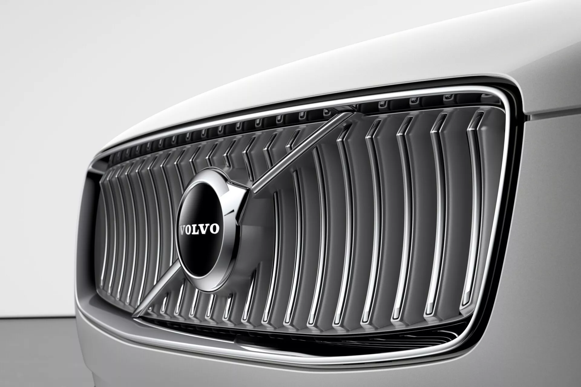 Vista en primer plano de la parrilla delantera de un Volvo XC90 2015 (2da Generación) desde un ángulo de tres cuartos delantero, destacando el emblema prominente de Volvo centrado en las lamas verticales cromadas, bajo una iluminación suave que resalta la pintura del coche de color claro, probablemente blanco o gris muy claro, y el elegante y moderno diseño cóncavo de la parrilla.