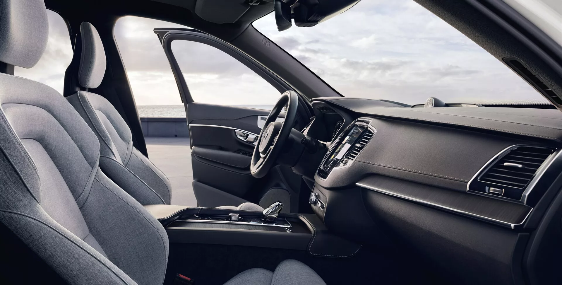 Vista interior de un Volvo XC90 2015 (2ª generación) desde la perspectiva del pasajero delantero, mostrando el asiento ergonómico del conductor tapizado en tela premium oscura o cuero sintético, volante multifuncional con controles integrados, tablero elegante con el sistema de infoentretenimiento táctil apagado, ventilaciones de aire integradas, controles mínimos de climatización, consola central con almacenamiento, portavasos y palanca de cambios, todo resaltado en un esquema de color oscuro unificado con luz natural iluminando el vehículo.