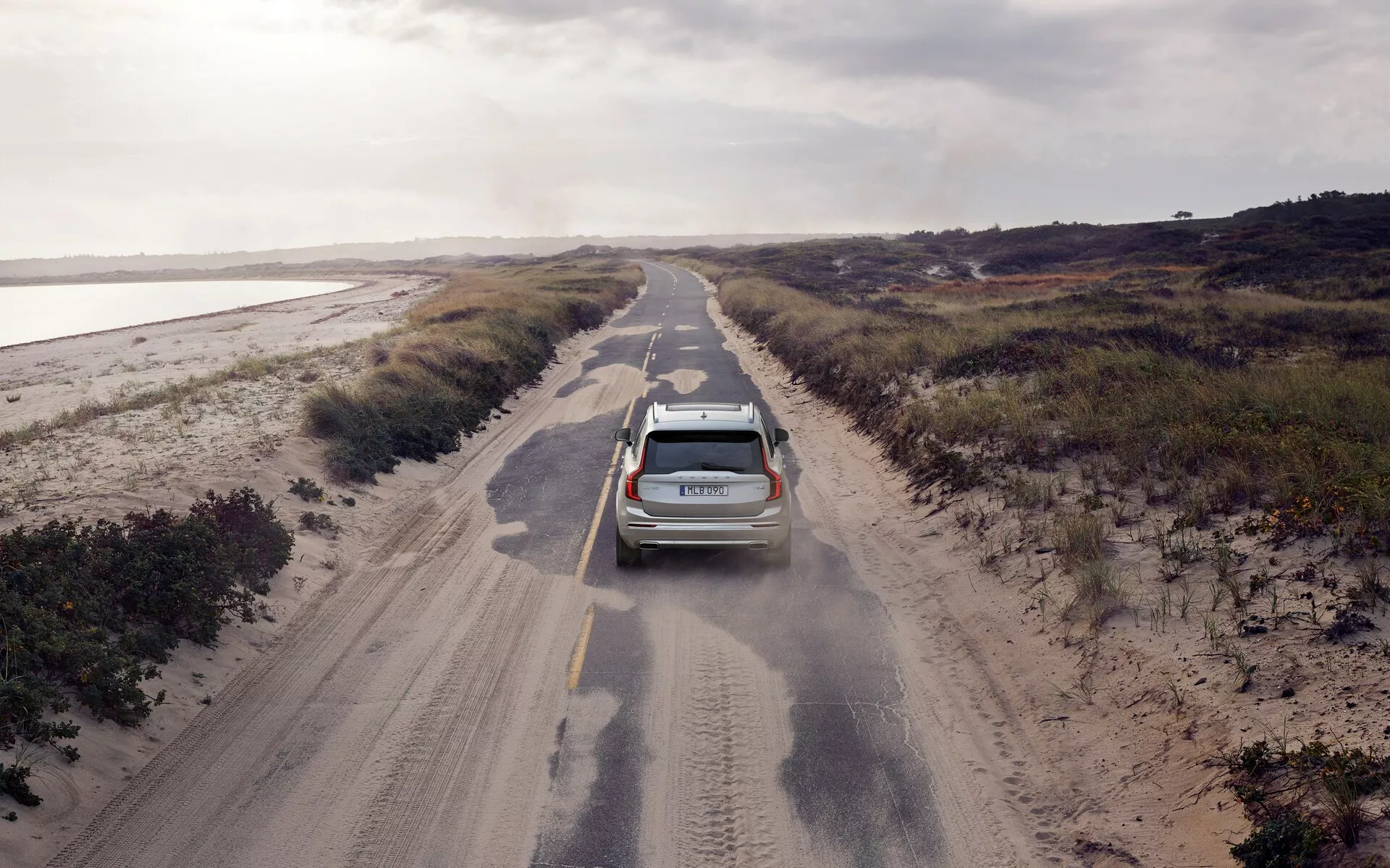 Vista aérea de un SUV Volvo XC90 2015 (2.ª generación) conduciendo por un camino estrecho y sin pavimentar, flanqueado por vegetación baja. El vehículo moderno, equipado con una antena y barras de techo, está ligeramente descentrado a la izquierda y levanta polvo, sugiriendo movimiento y la textura rugosa del camino. La iluminación nublada ilumina uniformemente la escena, contribuyendo a una sensación de aislamiento. Las características visibles incluyen luces traseras, un limpiaparabrisas trasero y posiblemente sensores de estacionamiento.
