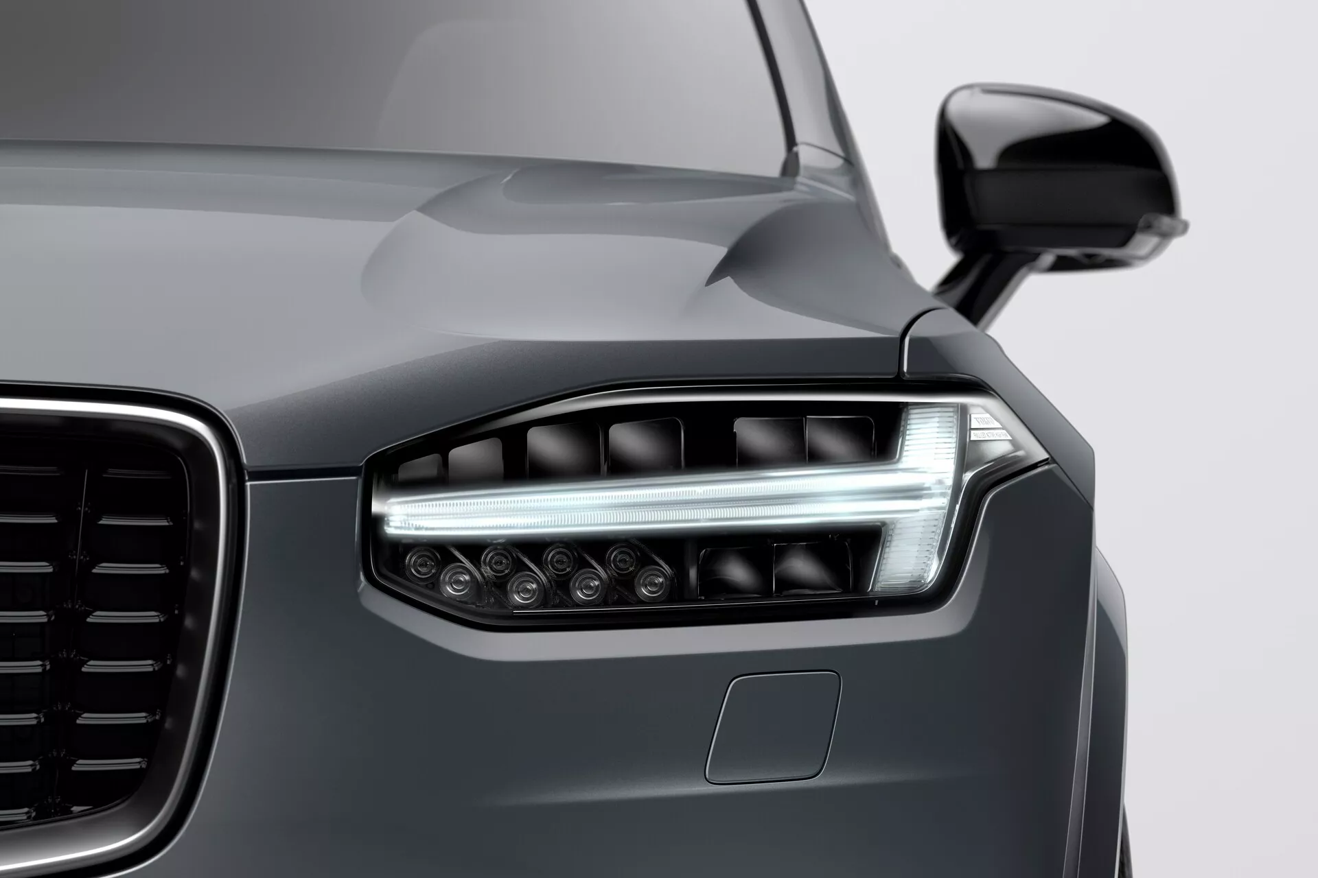 Vista en primer plano de la esquina delantera izquierda de un Volvo XC90 2015 (2ª generación), mostrando el diseño moderno de los faros LED con luces diurnas angulares, una vista parcial de una parrilla grande con patrón vertical, pintura metálica gris oscuro y un sensor de estacionamiento en el parachoques, tomada desde un ángulo bajo de derecha a izquierda.