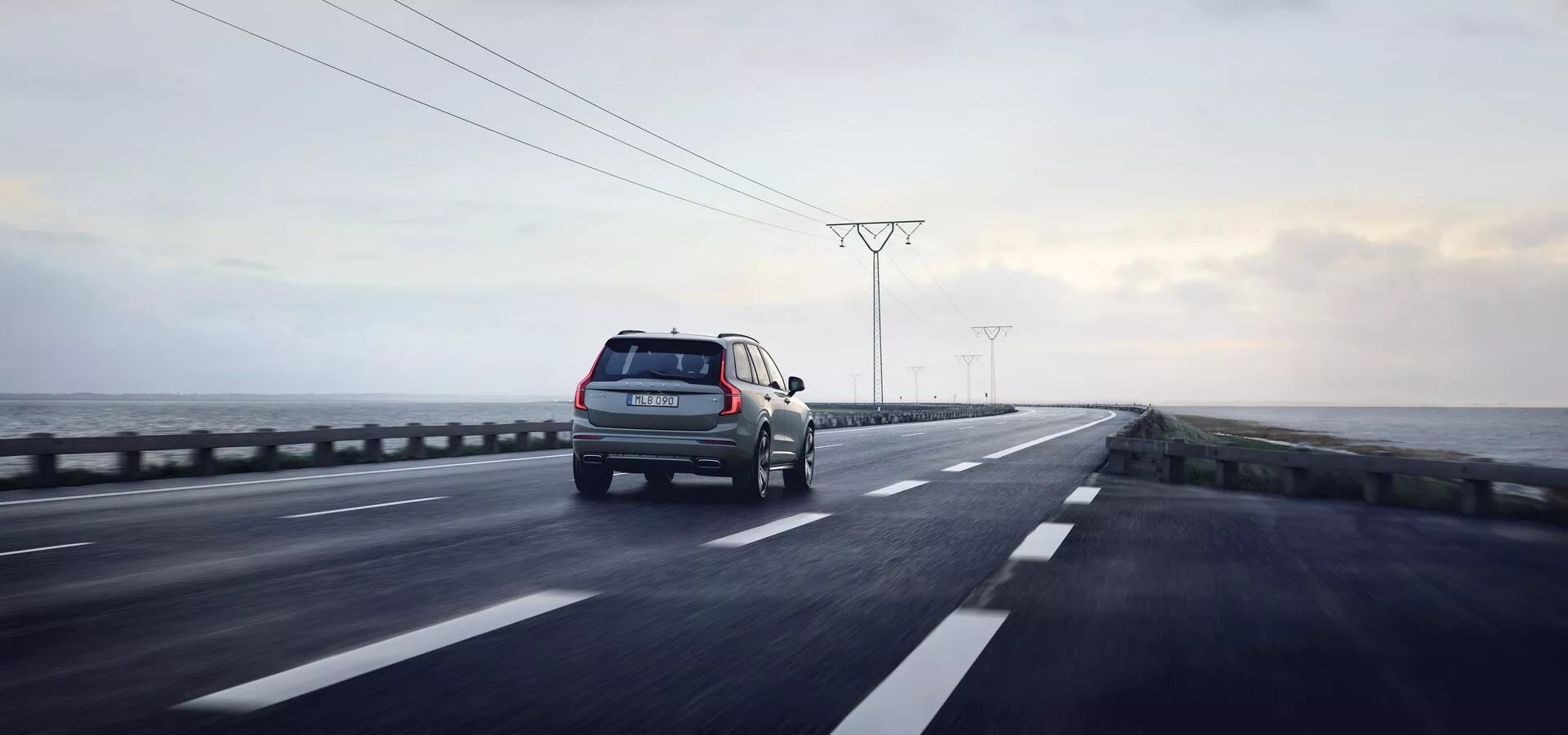 Vista trasera tres cuartos de un SUV en movimiento en una carretera de doble calzada con sistema de conducción a la derecha. El vehículo, situado en el carril derecho, probablemente sea un SUV dado su mayor altura y proporciones cuadradas. Características visibles incluyen luces traseras, vista parcial del lado derecho con puerta trasera y neumático, portaequipajes, ruedas en movimiento, parachoques trasero y reflectores. La carretera está húmeda con reflejos que indican un acabado brillante del coche. La escena circundante consiste en un cielo nublado, paisaje desolado con postes eléctricos, sugiriendo un entorno reciente de lluvia. La toma captura tanto el coche como el fondo en un amplio encuadre con profundidad de campo, evocando temas de tecnología, viajes y actividades al aire libre con iluminación natural atenuada.