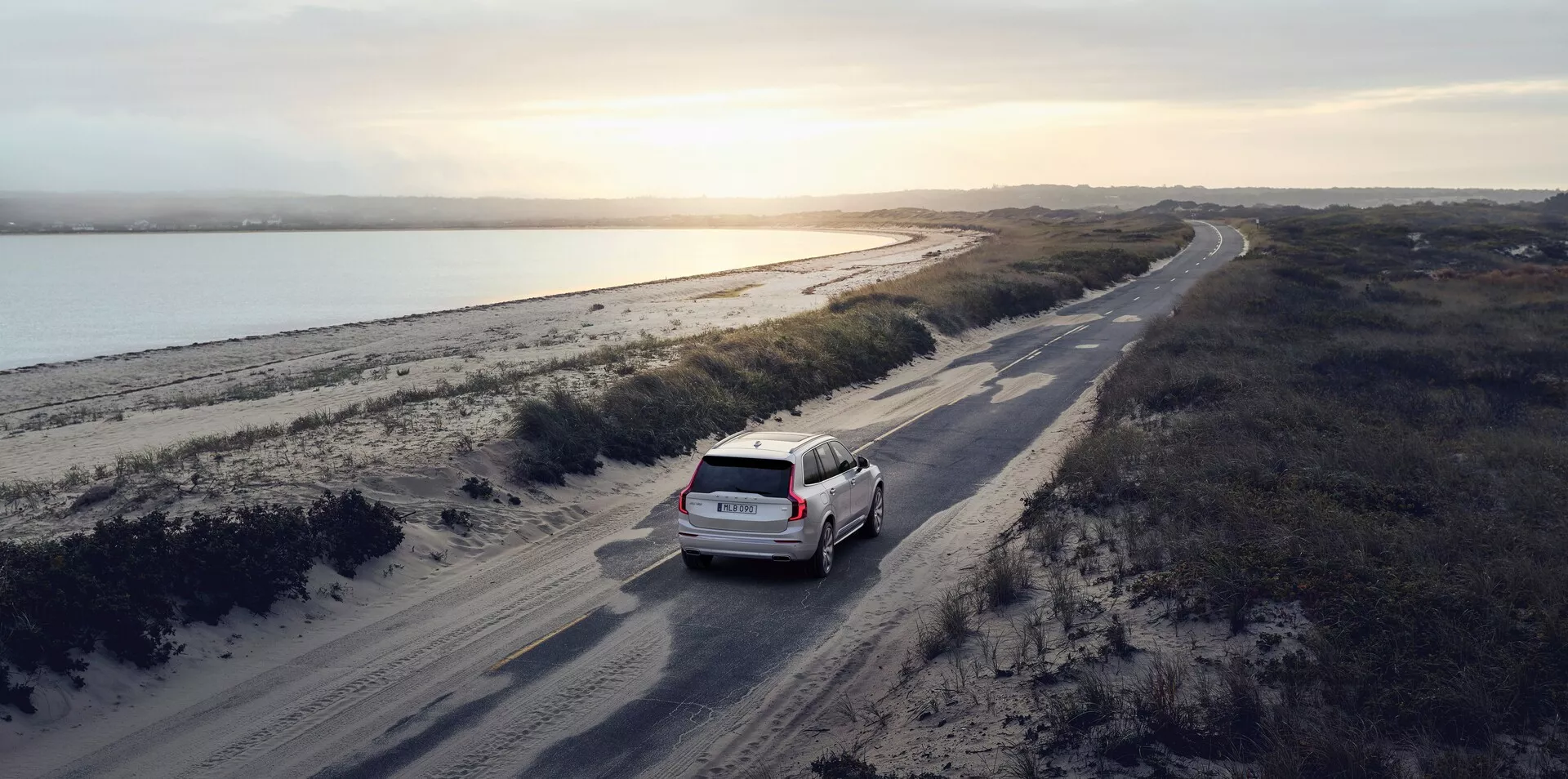 Toma aérea de un SUV Volvo XC90 2015 (2ª generación) blanco o de color claro conduciendo en un camino de tierra sinuoso paralelo a un cuerpo de agua, posiblemente un río, con un paisaje costero a la derecha. La escena ocurre al amanecer o al atardecer con luz natural suave que proyecta largas sombras, realzando los detalles del coche y su entorno. El coche está ligeramente a la derecha del centro en el tercio inferior de la imagen, mostrando su parte trasera, incluyendo la puerta trasera y el parabrisas trasero. La imagen enfatiza un sentido de aventura y viaje en un entorno natural pintoresco.