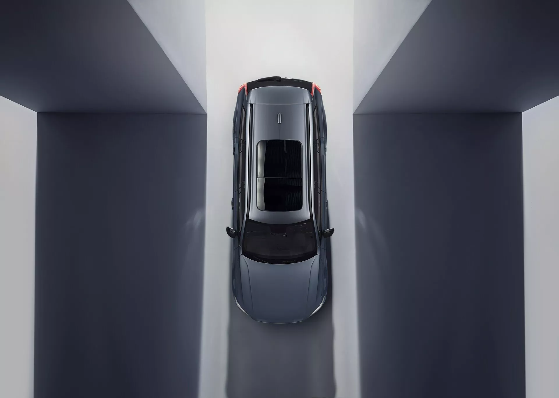 Vista aérea de un Volvo XC90 2015 (2ª generación) que muestra una composición simétrica y reflejos ambientales. La posición central en un entorno interior de tonos neutros resalta el techo del coche con un techo solar o techo panorámico de cristal. Las partes visibles incluyen el capó, el techo, el maletero y la mitad superior de las puertas, enfatizando líneas y curvas elegantes que sugieren un diseño lujoso centrado en la aerodinámica y una iluminación interior espaciosa.