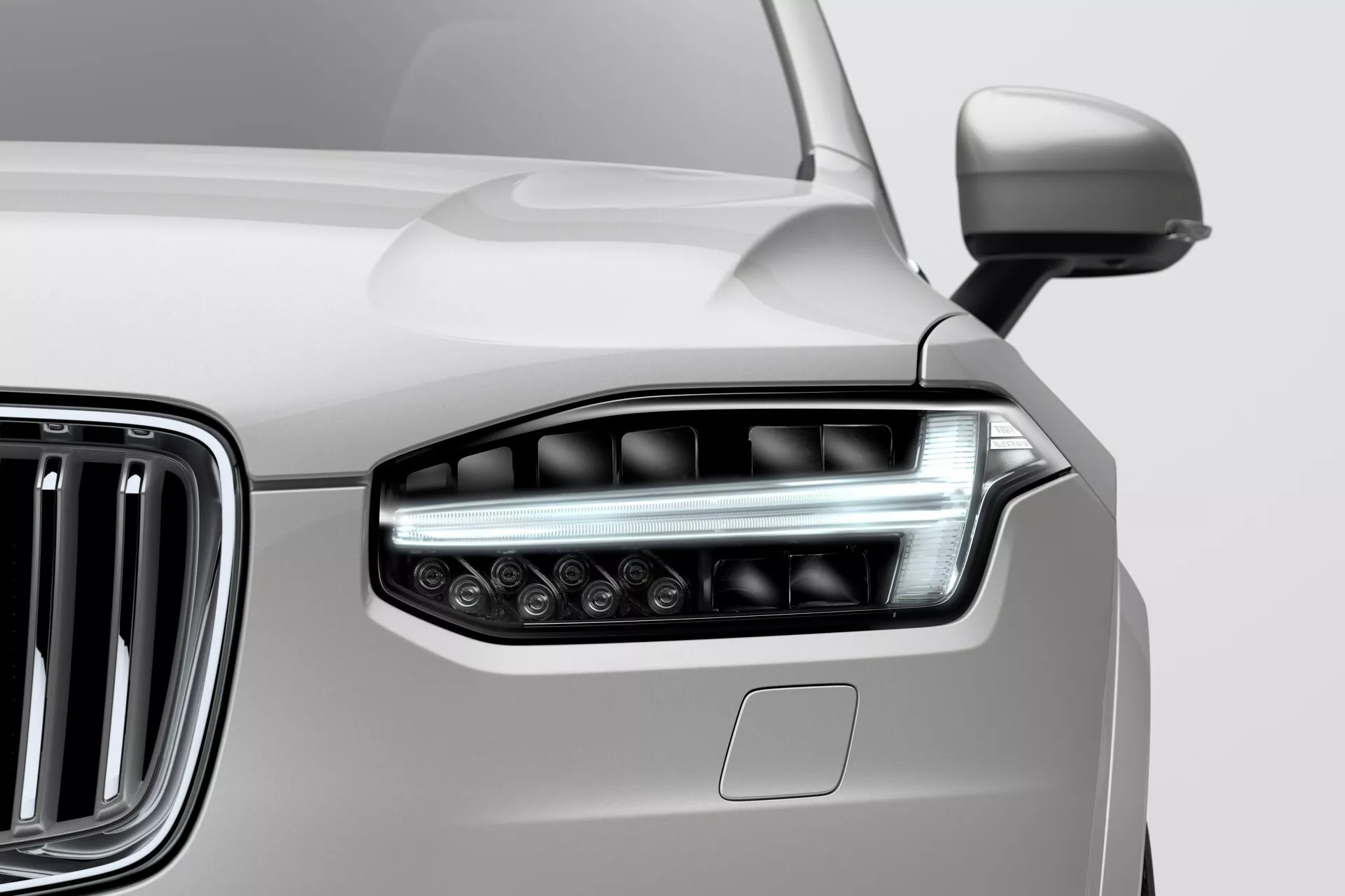 Vista en primer plano del conjunto del faro delantero izquierdo y parte de la parrilla de un Volvo XC90 2015 (segunda generación). El faro elegante integra elementos de iluminación LED/HID avanzados y alargados con una lente clara, presentando un diseño angular y aerodinámico. La parrilla cromada adyacente con listones verticales resalta la apariencia moderna y lujosa del vehículo. El panel de la carrocería, probablemente de color neutro como plateado, muestra una transición suave desde el faro. También se vislumbra parcialmente un espejo lateral. La imagen enfatiza el estilo contemporáneo del coche y su avanzada tecnología de iluminación.