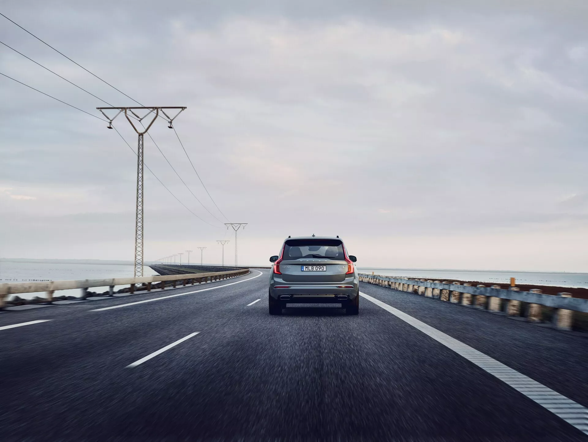 Vista trasera de un Volvo XC90 2015 (2ª generación) viajando por una carretera costera de dos carriles. La foto está tomada desde un ángulo bajo y una perspectiva centrada, capturando la tapa del maletero, el parabrisas trasero, las luces traseras, el parachoques trasero y el distintivo de marca y modelo. El vehículo es el principal enfoque, con un horizonte oceánico ligeramente borroso y torres eléctricas que se desvanecen en la distancia bajo un cielo gris y nublado. La matrícula del coche es visible y la iluminación sugiere temprano en la mañana o tarde en la tarde con una luz difusa.