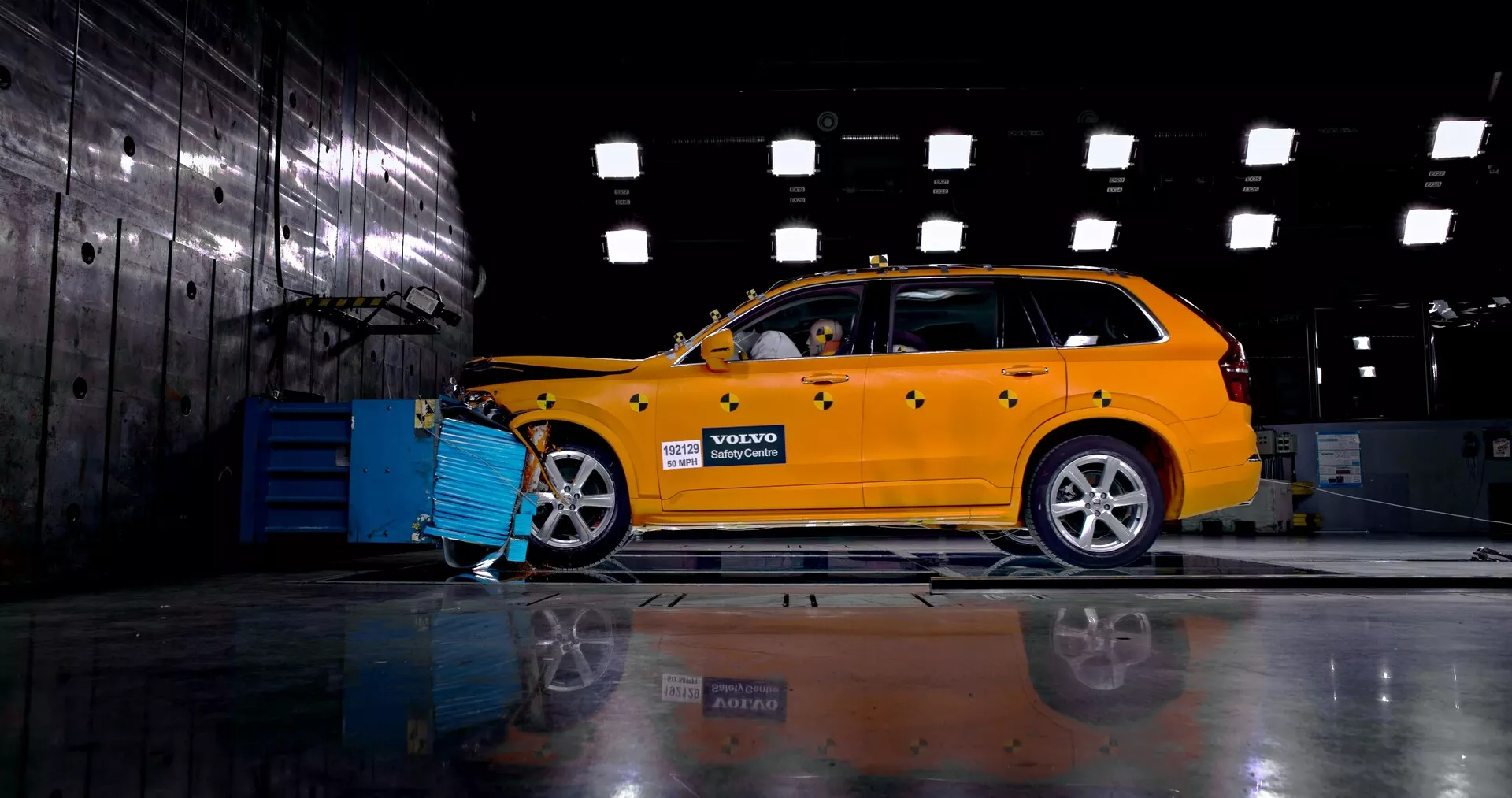 Vista de perfil lateral de un Volvo XC90 2015 (2ª generación) de color amarillo brillante en un entorno industrial, mostrando su diseño de carrocería familiar con parrilla delantera visible, faros y ruedas de aleación. El coche está conectado a un equipo de diagnóstico o carga azul y un maniquí dentro sugiere una demostración de prueba de seguridad o choque. El suelo pulido refleja el chasis del coche y las ruedas del lado opuesto.