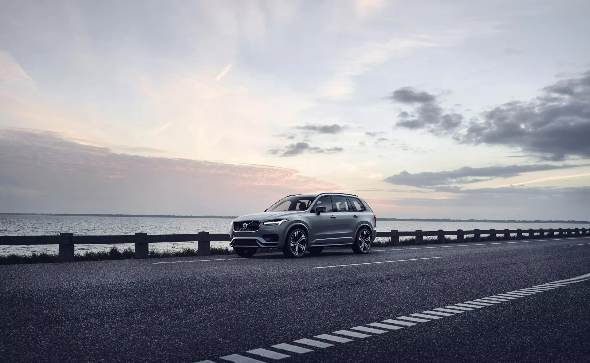 Un SUV moderno, específicamente el Volvo XC90 2015 (2ª generación), estacionado en el lado derecho de una carretera de dos carriles. El vehículo está capturado desde una vista frontal de tres cuartos, mostrando la parrilla delantera, los faros, el capó y el guardabarros del lado del conductor. El ángulo también revela parte del perfil del coche, incluyendo las ruedas, la puerta del conductor y parte de la parte trasera. Las ruedas están ligeramente giradas a la derecha, con elementos de diseño posiblemente deportivos como un parachoques pronunciado y tomas de aire más grandes que sugieren un paquete dinámico. La foto, tomada probablemente al atardecer con luz suave y difusa bajo un cielo ligeramente nublado, realza el ambiente sereno. La carrocería bien definida con líneas de carácter visible indica un modelo de gama alta. Las características tecnológicas inferidas de la imagen incluyen faros LED, diseños avanzados de ruedas, una parrilla frontal sofisticada con posibles sensores o cámaras y toques aerodinámicos. Esta imagen de alta calidad podría ser adecuada para publicidad, enfatizando el coche como una elección de estilo de vida lujoso.