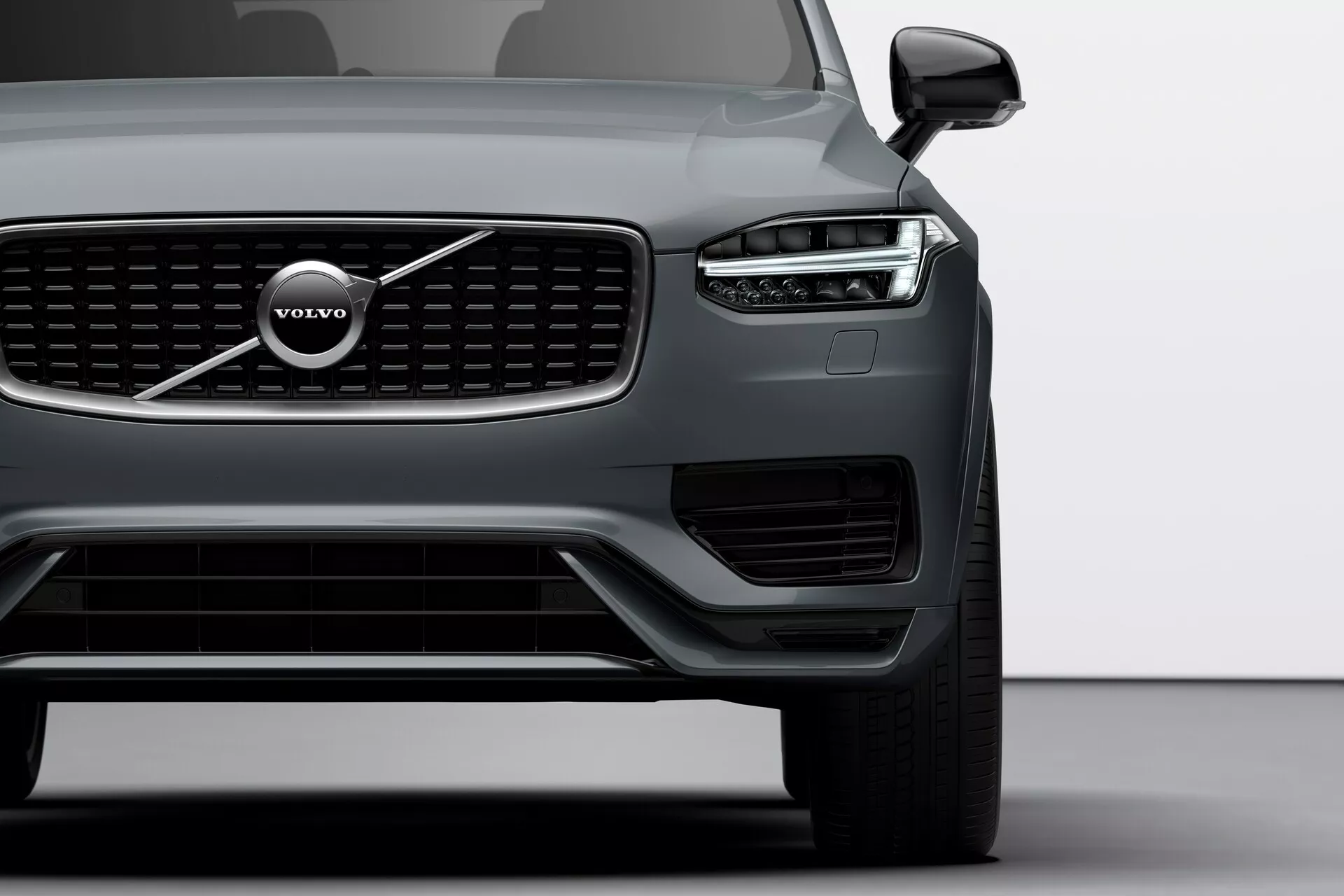Vista del cuarto delantero izquierdo de un Volvo XC90 2015 (2da generación), tomada desde una perspectiva de ángulo bajo que muestra los faros angulares y afilados con distintivas luces diurnas LED en forma de martillo de Thor, la parrilla delantera prominente con listones verticales con el emblema Volvo, la amplia entrada de aire en el parachoques inferior con luces/sensores adicionales, los contornos aerodinámicos del capó, el espejo exterior elegante con señal de giro integrada, la rueda de aleación grande de múltiples radios parcialmente visible y el acabado mate de la pintura del cuerpo, destacando el diseño modern y avanzada tecnología del vehículo.