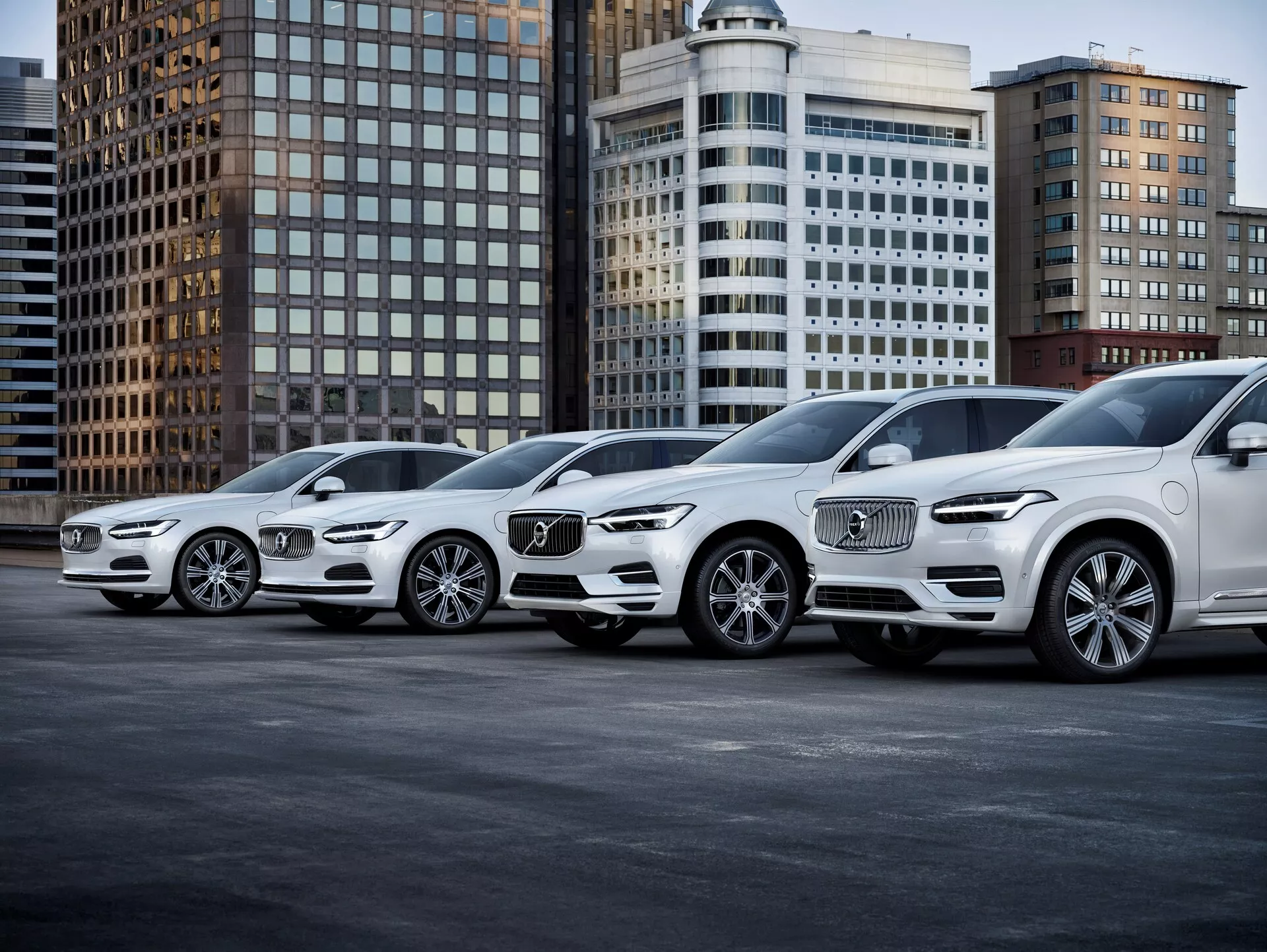 La imagen muestra una alineación de cuatro lujosos SUVs Volvo XC90 de segunda generación de 2015, inclinados con sus perfiles frontales orientados hacia la derecha. Colocados en diagonal, la perspectiva resalta tanto las vistas frontales como laterales de los vehículos. Los coches presentan rejillas delanteras prominentes con el reconocible logo de Volvo, faros LED intrincados, diseños de ruedas de aleación y líneas de contorno en el capó. Capturada desde un ángulo bajo cerca del suelo, la fotografía enfatiza el tamaño y la postura de los SUVs, ubicados en un fondo urbano con edificios de oficinas. La iluminación es suave, lo que sugiere un día nublado o un entorno urbano bien iluminado, mostrando claramente las ruedas, faros, luces antiniebla y líneas de diseño. El vehículo más cercano muestra tecnologías de iluminación avanzadas y sensores de rejilla delanteros, probablemente asociados con conducción autónoma o asistencia en el estacionamiento. A medida que los coches se alejan, su estilo idéntico indica que pertenecen a la misma familia de vehículos, lo que hace que esta imagen sea ideal para las categorías de Automoción, Vehículos de Lujo, Tecnologías Avanzadas de Vehículos y Estilo de Vida Urbano.