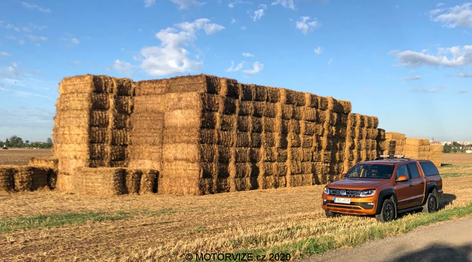 Un Volkswagen Amarok 2016 estacionado frente a grandes fardos de heno en un entorno rural. La perspectiva frontal de tres cuartos captura la parrilla del coche, los faros, el parachoques delantero, el capó, las ruedas, los espejos laterales y las puertas. La prominente parrilla de listones horizontales y los faros angulares agudos realzan el aspecto contemporáneo del vehículo. Las ventanas tintadas, los rieles del techo y el revestimiento protector alrededor de las puertas sugieren versatilidad y capacidad resistente. El vehículo está situado contra un fondo de fardos de heno, añadiendo contexto para su uso en conducción por el campo o en granjas. La luz de la mañana o del final de la tarde proyecta sombras cálidas y realza el color y los detalles de diseño del coche. Con marca de agua 'MOTORVIZE.cz 2020'.