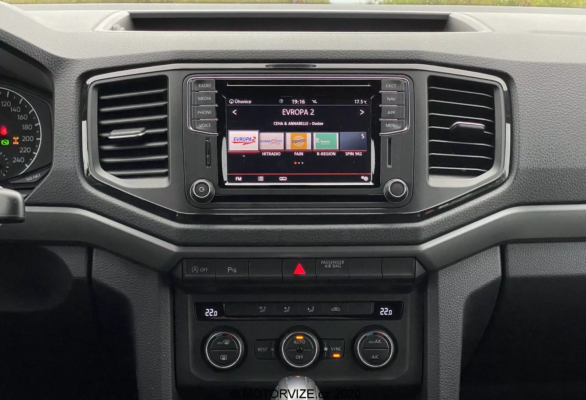 La imagen presenta el tablero central del VW Nutzfahrzeuge Amarok 2016. Desde una perspectiva frontal, se puede ver el sistema de infoentretenimiento mostrando una estación de radio, dos ventilas de aire horizontales y varios controles. El sistema de audio y el control climático, incluidos los ajustes de temperatura y ventilador, son claramente visibles. El tablero prioriza la funcionalidad con botones y perillas fáciles de usar, utilizando principalmente materiales plásticos duros, adecuados para el uso diario en lugar de lujo.