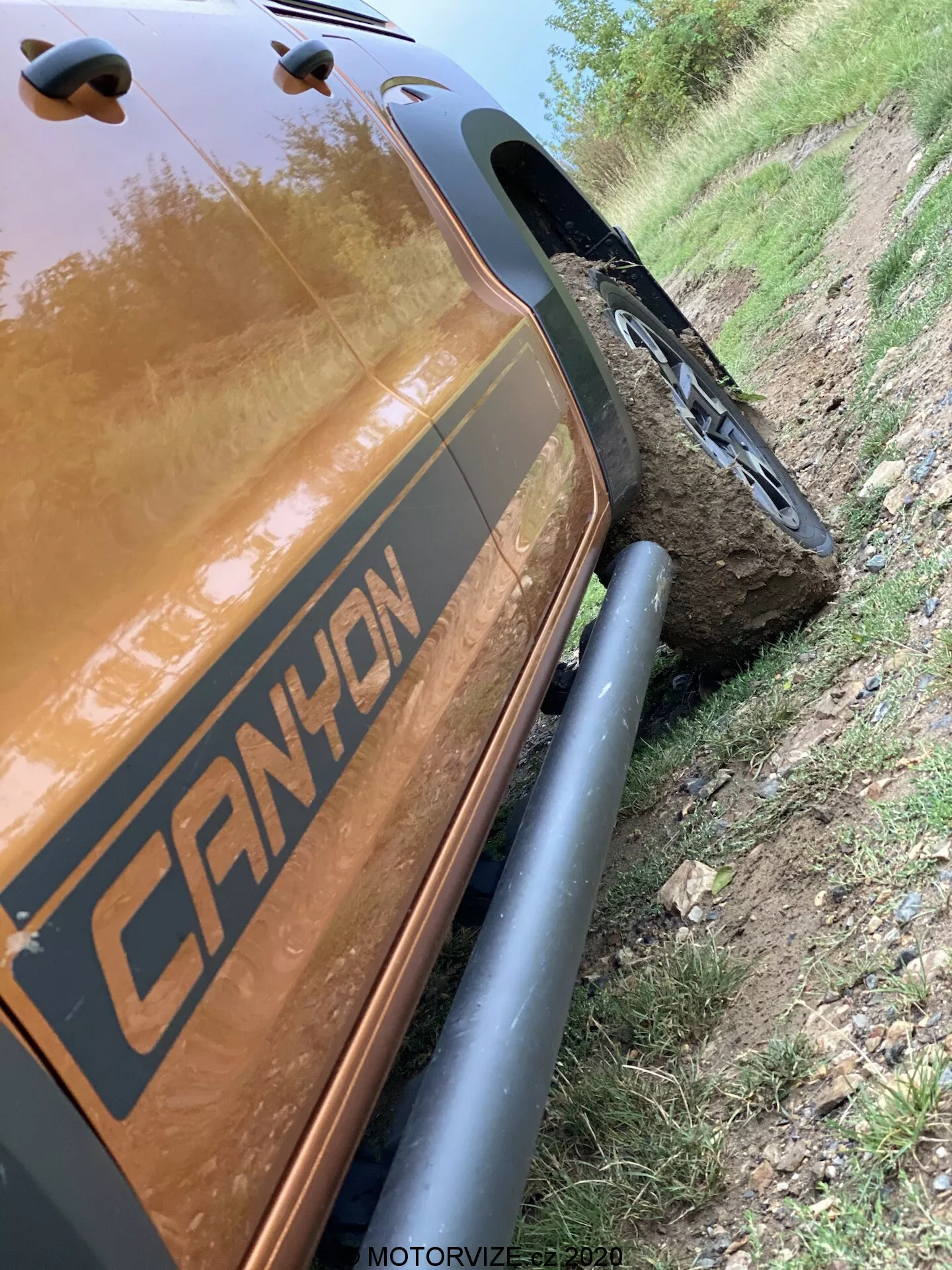 Esta imagen con ángulo inclinado de 45 grados muestra la parte trasera del lado del pasajero de un VW-Nutzfahrzeuge Amarok 2016. Enfoca los detalles cercanos desde el paso de rueda trasero hasta la sección de carga trasera, destacando los robustos protectores negros del arco de la rueda y los neumáticos todo terreno rugosos. El vehículo presenta una gráfica con la leyenda 'CANYON' y está ubicado junto a un camino de tierra con una rueda montada en una gran piedra, demostrando su capacidad todoterreno. El color metálico reflectante marrón o cobre del cuerpo del automóvil es evidente, capturando reflejos de luz. La marca de tiempo 'MOTORVIZE.cz 2020' sugiere que fue capturado para una publicación de vehículos a motor.