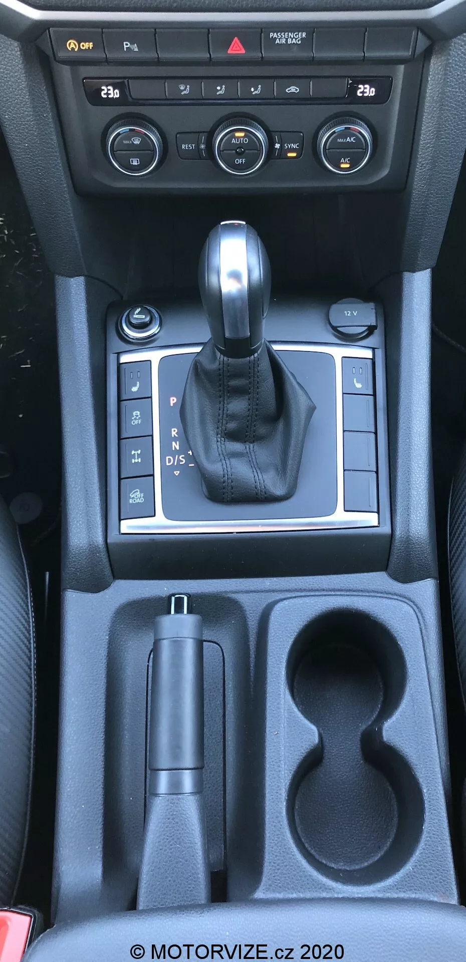 Esta imagen muestra la consola central de un vw-nutzfahrzeuge amarok 2016, tomada desde una perspectiva aérea aproximadamente desde el punto de vista del conductor o el pasajero delantero. La fotografía captura el interior del coche especificando la palanca de cambios, los controles de la consola central y los compartimentos de almacenamiento. La imagen incluye diales y botones del sistema de control de clima, una palanca de cambios revestida en cuero en un panel plateado que indica transmisión automática, un control de freno de estacionamiento electrónico, un compartimento de almacenamiento, dos portavasos y un almacenamiento adicional hacia los asientos traseros. Los materiales visibles incluyen plástico, acentos metálicos y cuero, reflejando una mezcla de funcionalidad y estética.