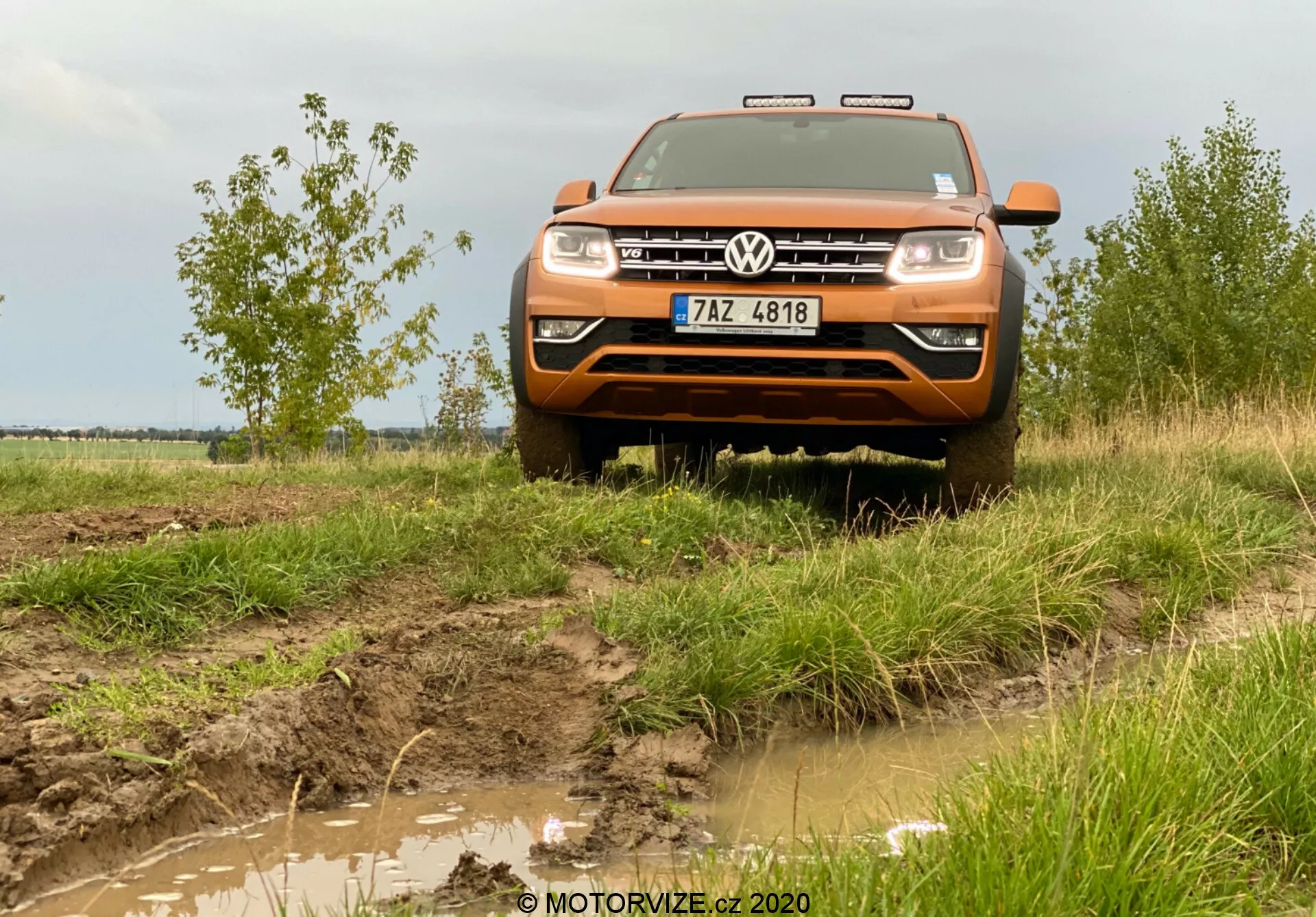 Vista frontal, ligeramente baja, de un Volkswagen Amarok 2016 naranja, una camioneta de tamaño mediano que muestra sus capacidades todoterreno. La imagen captua la parte delantera del vehículo, incluyendo el capó, la parrilla, el parachoques, parte de los guardabarros, los faros y el logotipo de Volkswagen en el centro de la parrilla. Sobre la parrilla, se ven acentos cromados o luces diurnas LED. El terreno fangoso y la hierba alrededor del automóvil enfatizan su naturaleza todoterreno, con luces adicionales en el techo y una placa de deslizamiento frontal con un telón de fondo rural de cielos nublados completando el entorno aventurero. La imagen se toma a una distancia de aproximadamente dos a tres metros, proporcionando una vista detallada de las características frontales del vehículo y su interacción con el entorno.